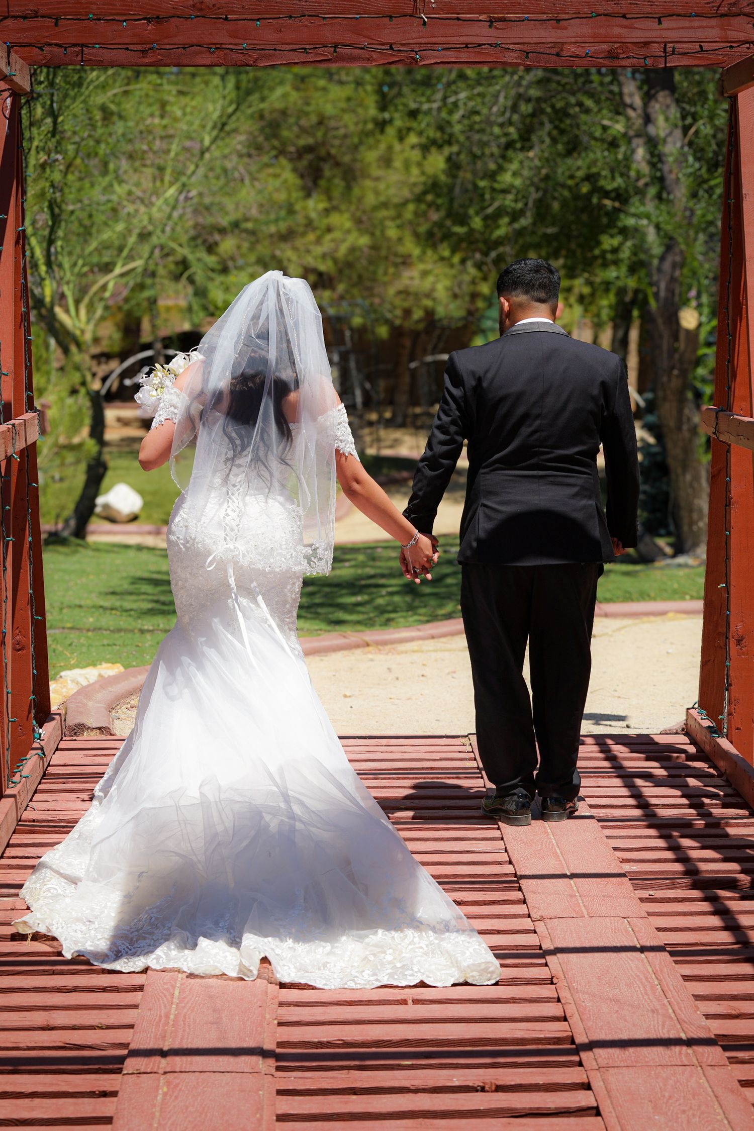 Newlywed couple walking hand-in-hand across a bridge after their outdoor wedding, seen from behind, with the bride’s long veil trailing.
