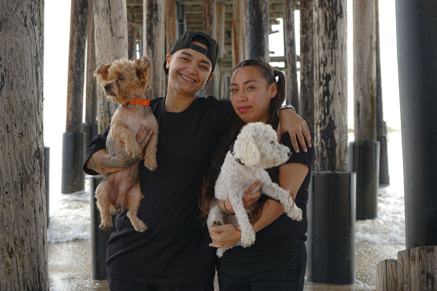 Couple embracing while holding their two small dogs, smiling, standing under a pier with the waves of the ocean receding behind them.