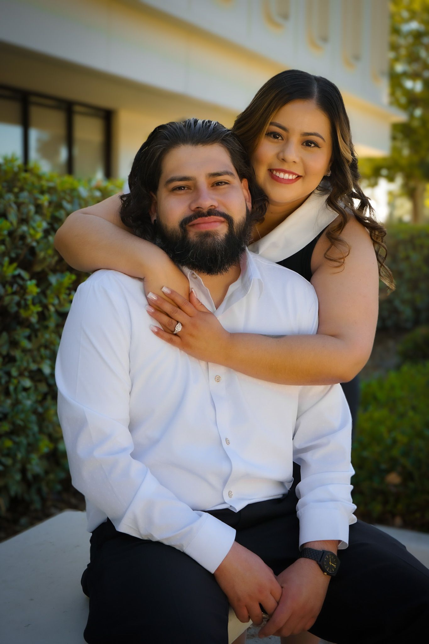 Engagement session for couple sitting on a bench outdoors, the bride to be is standing behind her fiancee, hugging him around the shoulders.