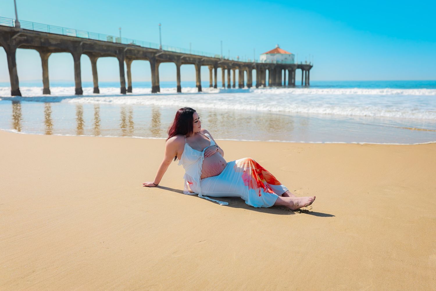 Pregnant mom seated on the sand in white and orange floral attire, gently cradling her belly with a serene expression.