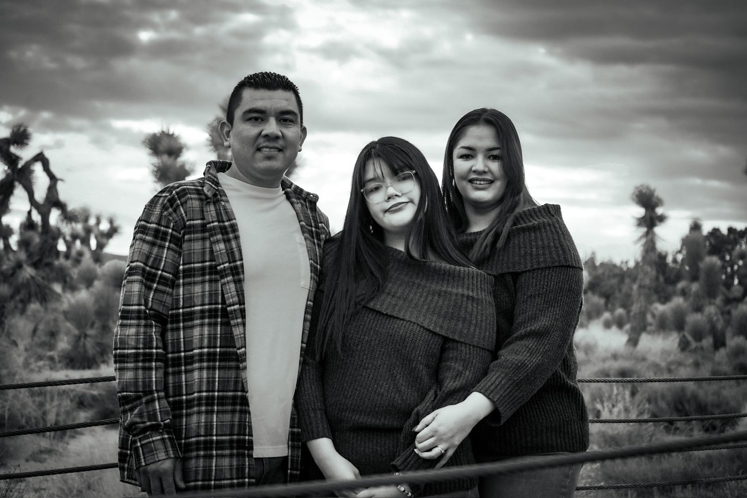 Family of three smiling warmly surrounded by desert foliage, a beautiful cloudy sky with hints of sunlight breaking through.