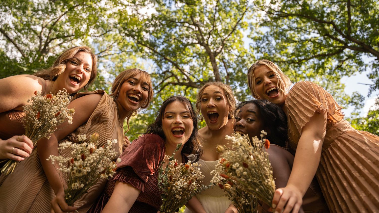 Group of friends laughing and celebrating outdoors under green trees.