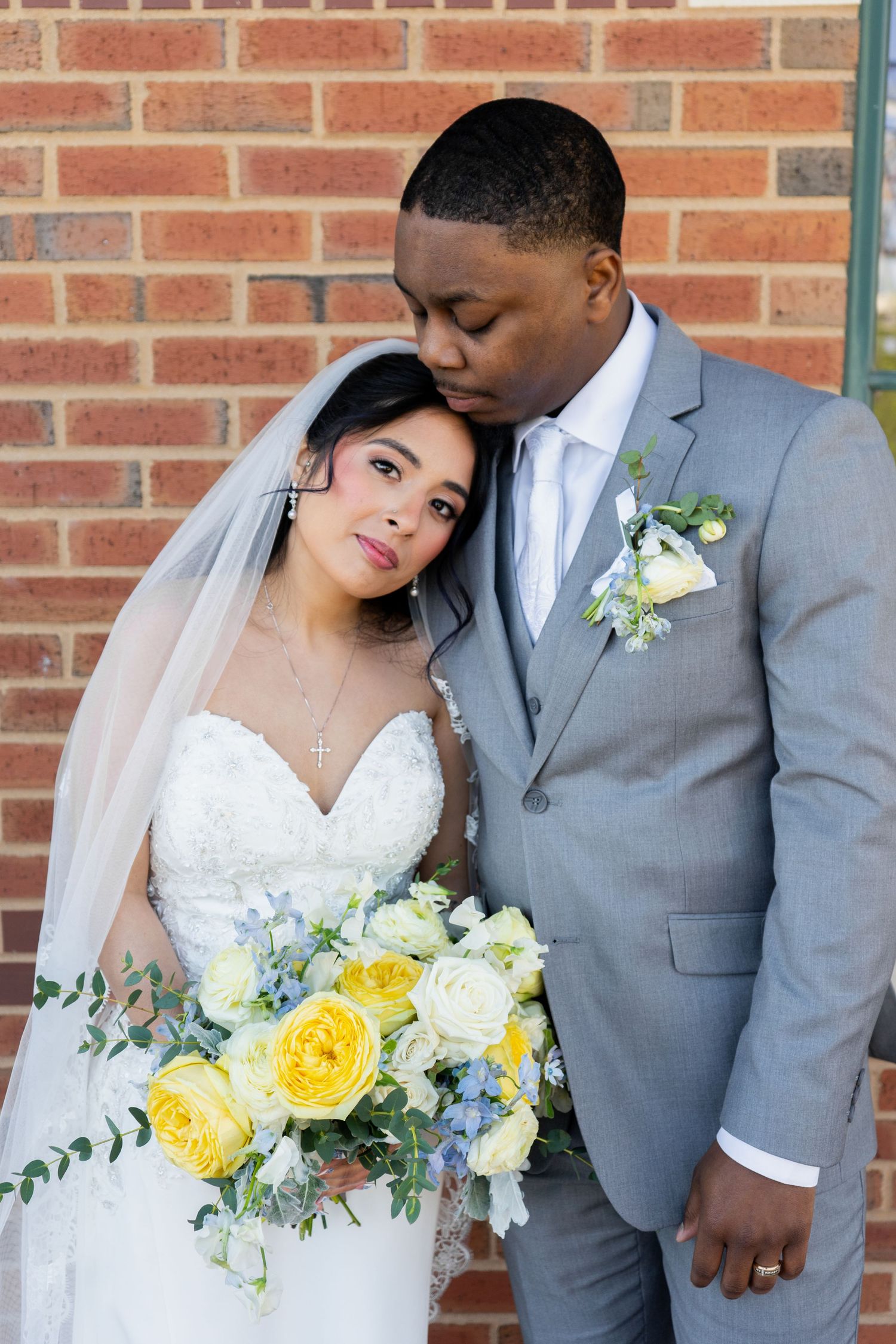 Bride and groom pose tenderly against brick wall, she holds yellow and white bouquet, he wears gray suit with boutonniere.