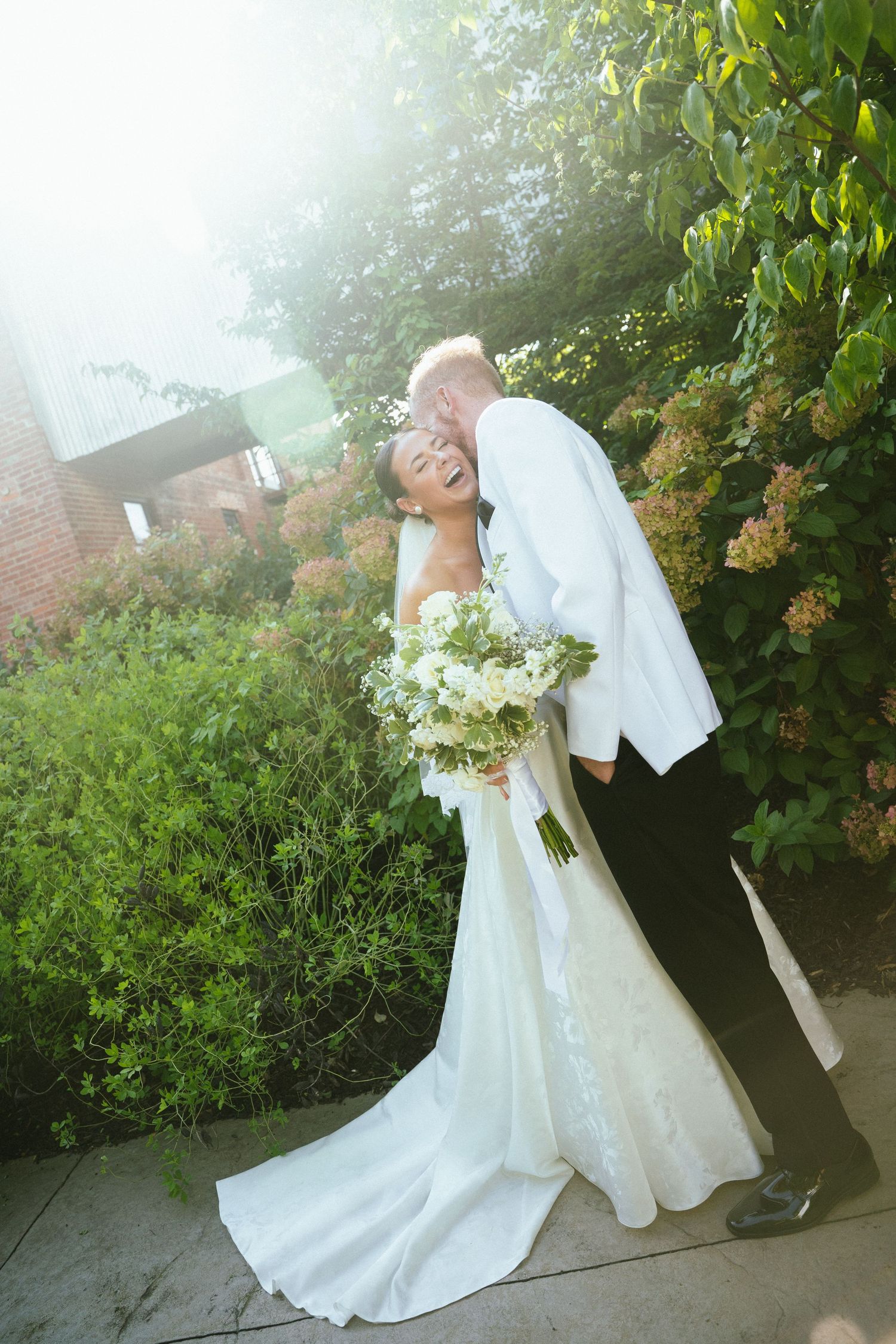 A groom in a white jacket kisses his laughing bride in a white gown holding a bouquet outdoors.