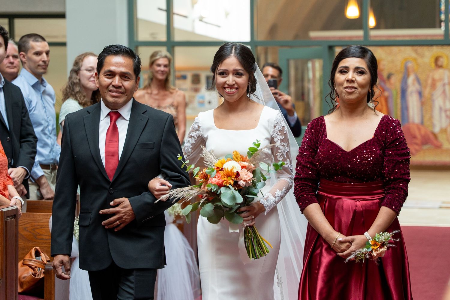 A joyful wedding processional inside a church with a bride in white carrying an orange and white bouquet.