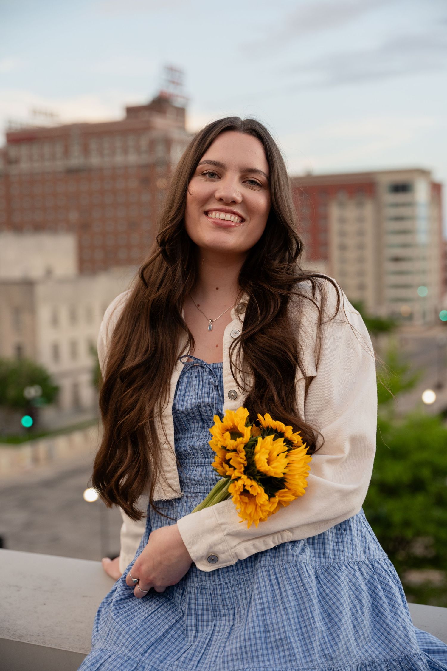 Person in blue gingham dress holds sunflowers against urban skyline backdrop.