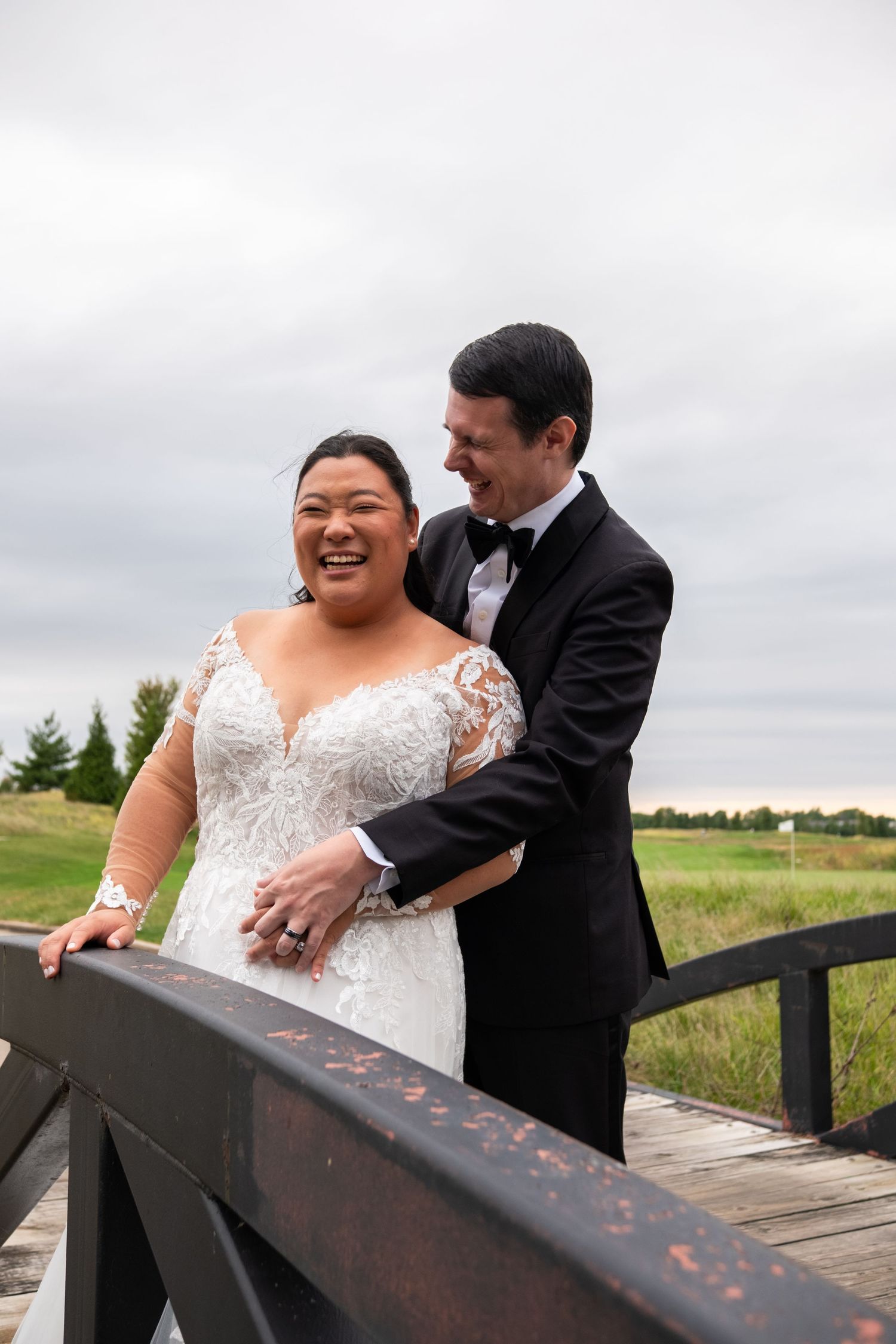 A joyful bride in lace gown laughs with her groom in a black tuxedo on a wooden bridge outdoors.