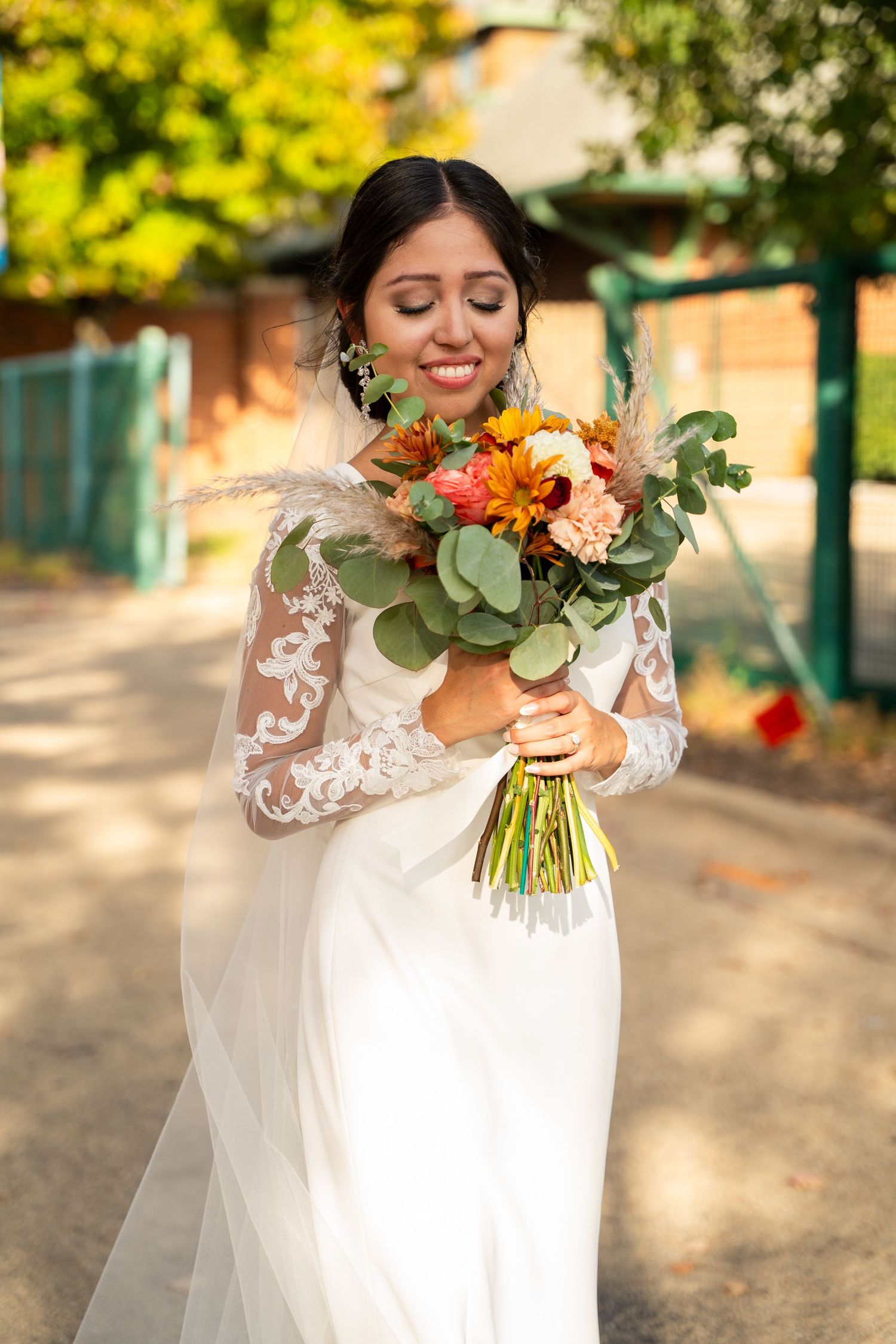 A bride in a white lace gown holds a vibrant autumn bouquet with orange and peach flowers in an outdoor setting.