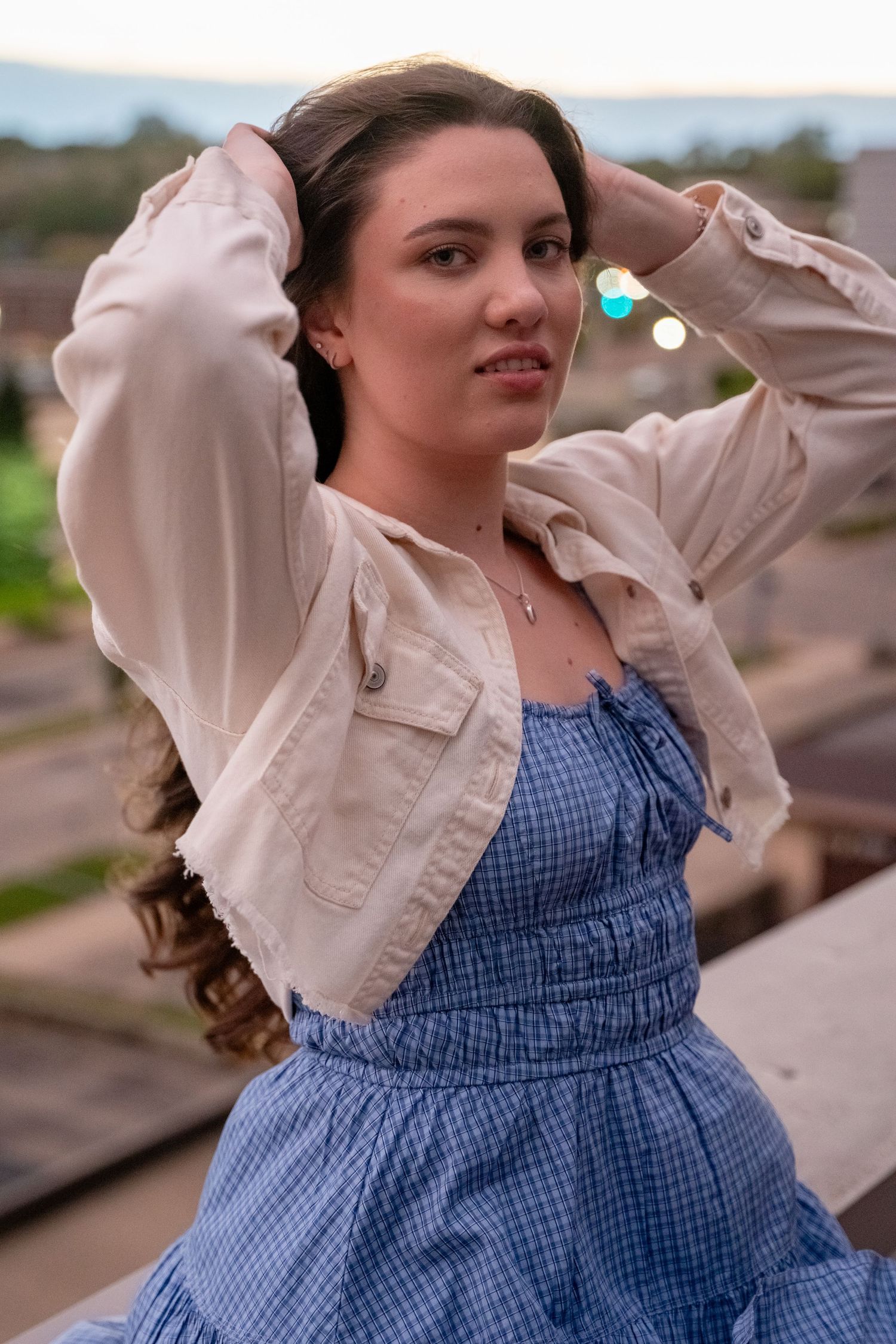 Person in blue gingham dress and cream jacket poses against sunset sky.