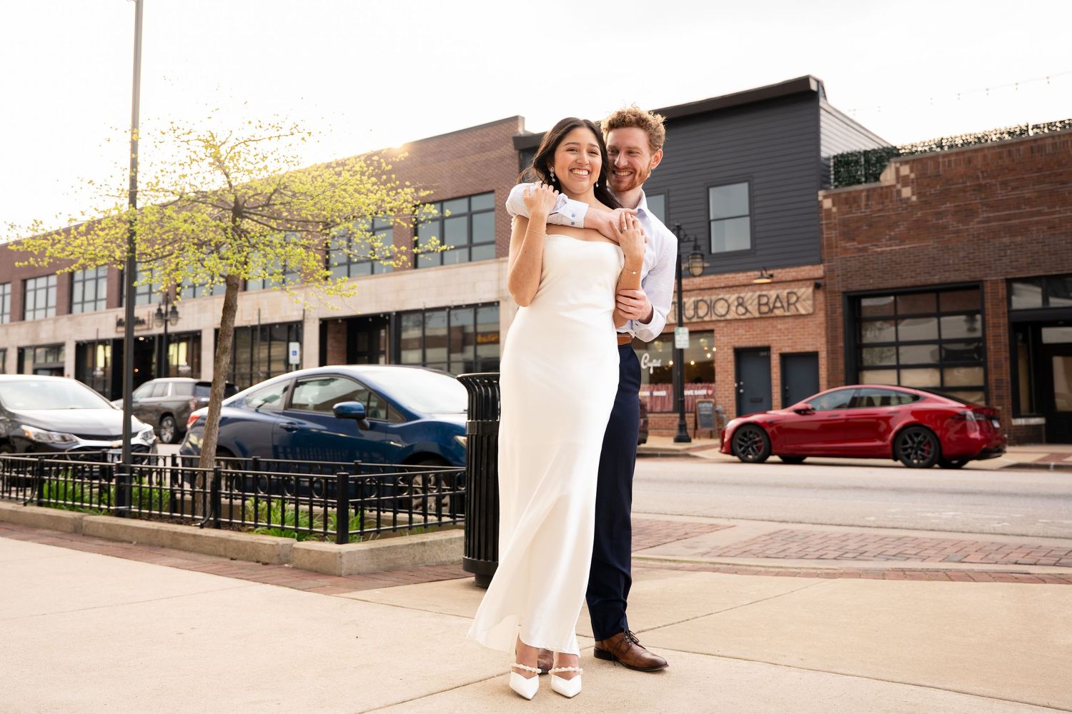 Wedding couple walking and dancing on urban sidewalk in front of brick buildings.