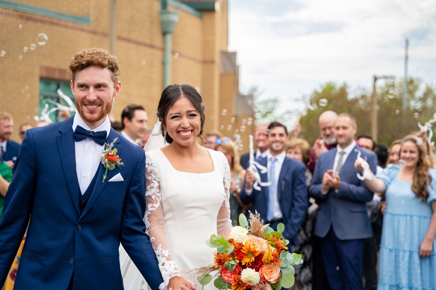 Wedding party celebrates outdoors with guests in navy blue and light blue attire and vibrant orange bouquets.