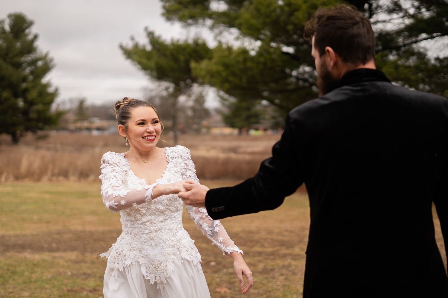 A smiling bride in a lace wedding dress holds hands with a groom in a black suit outdoors in a field.