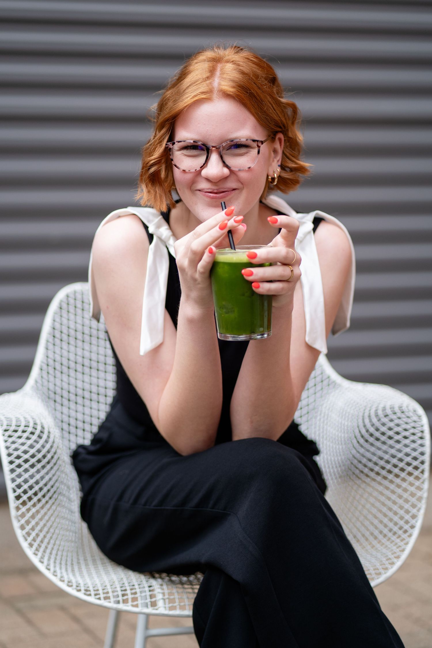 Person in black dress enjoys green smoothie while relaxing in white modern chair.