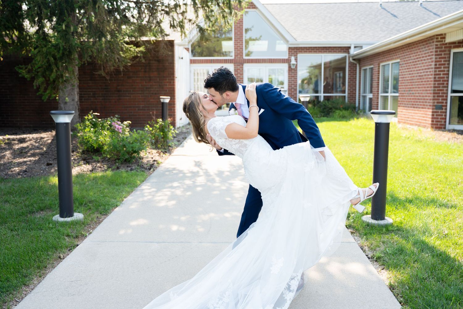 A couple shares a romantic dip kiss on a sunny sidewalk, with the bride's white dress flowing elegantly.