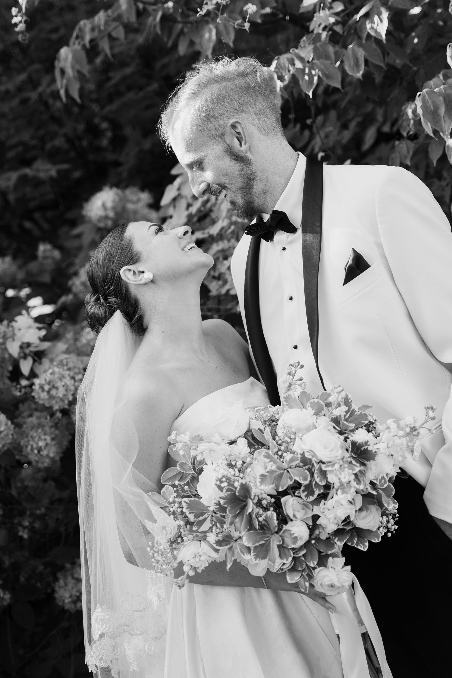 Black and white photo of a smiling bride and groom in formal wear sharing a romantic moment outdoors with floral bouquet.