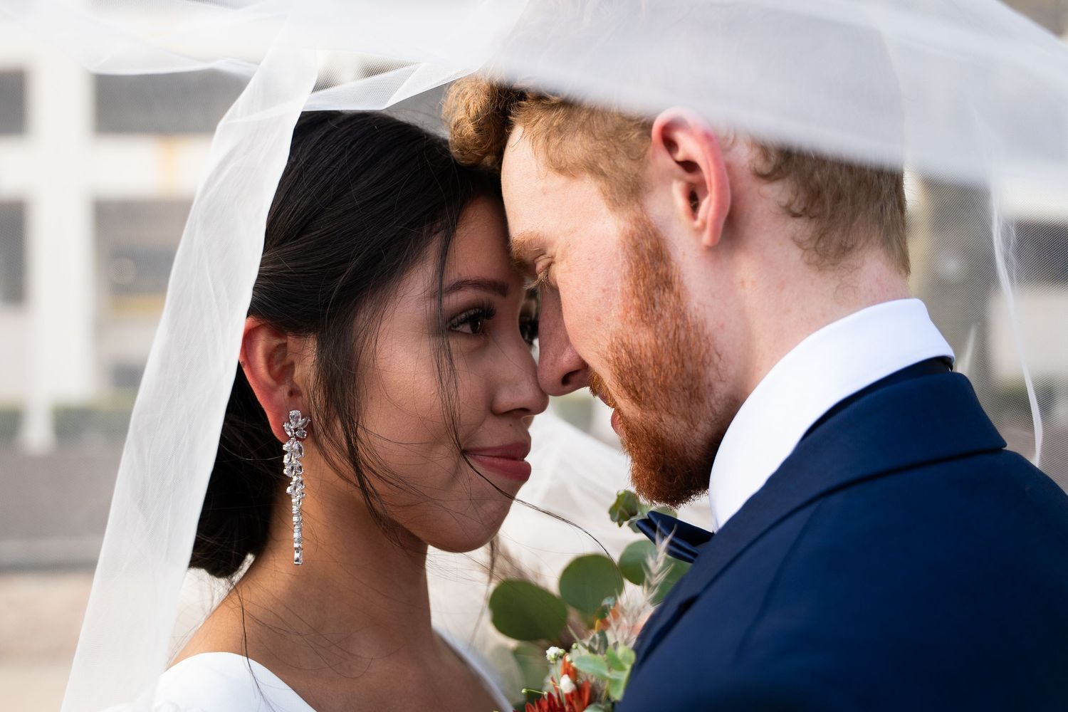 Bride and groom share an intimate moment under a wedding veil, touching foreheads in a romantic close-up portrait.