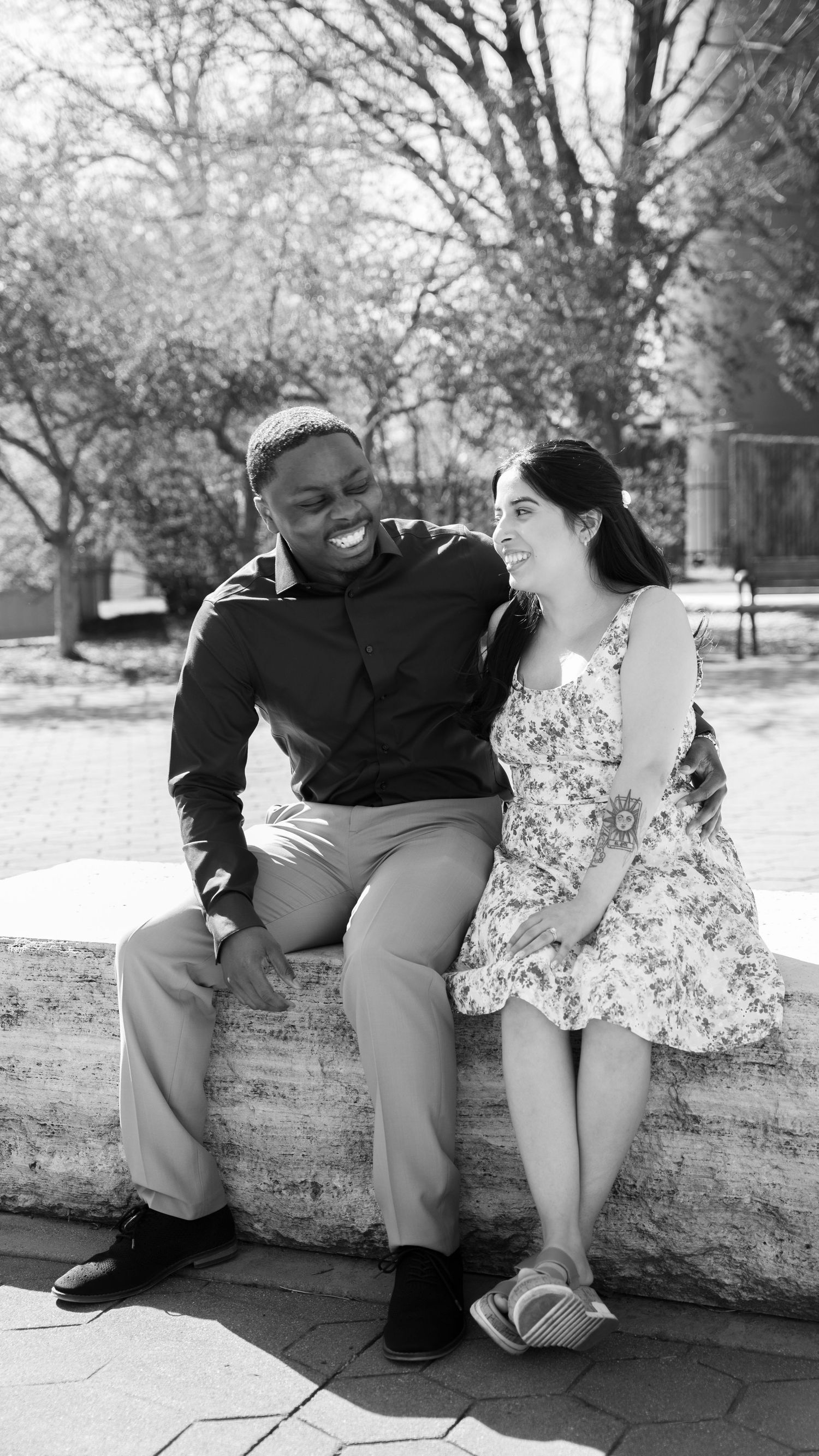 A couple sits together on a stone wall sharing a playful moment in black and white.
