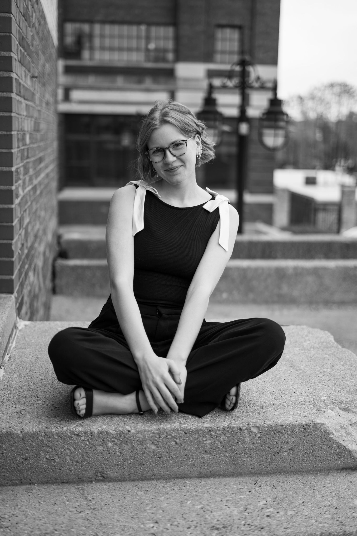 Person in black outfit sits casually on concrete ledge with urban backdrop.