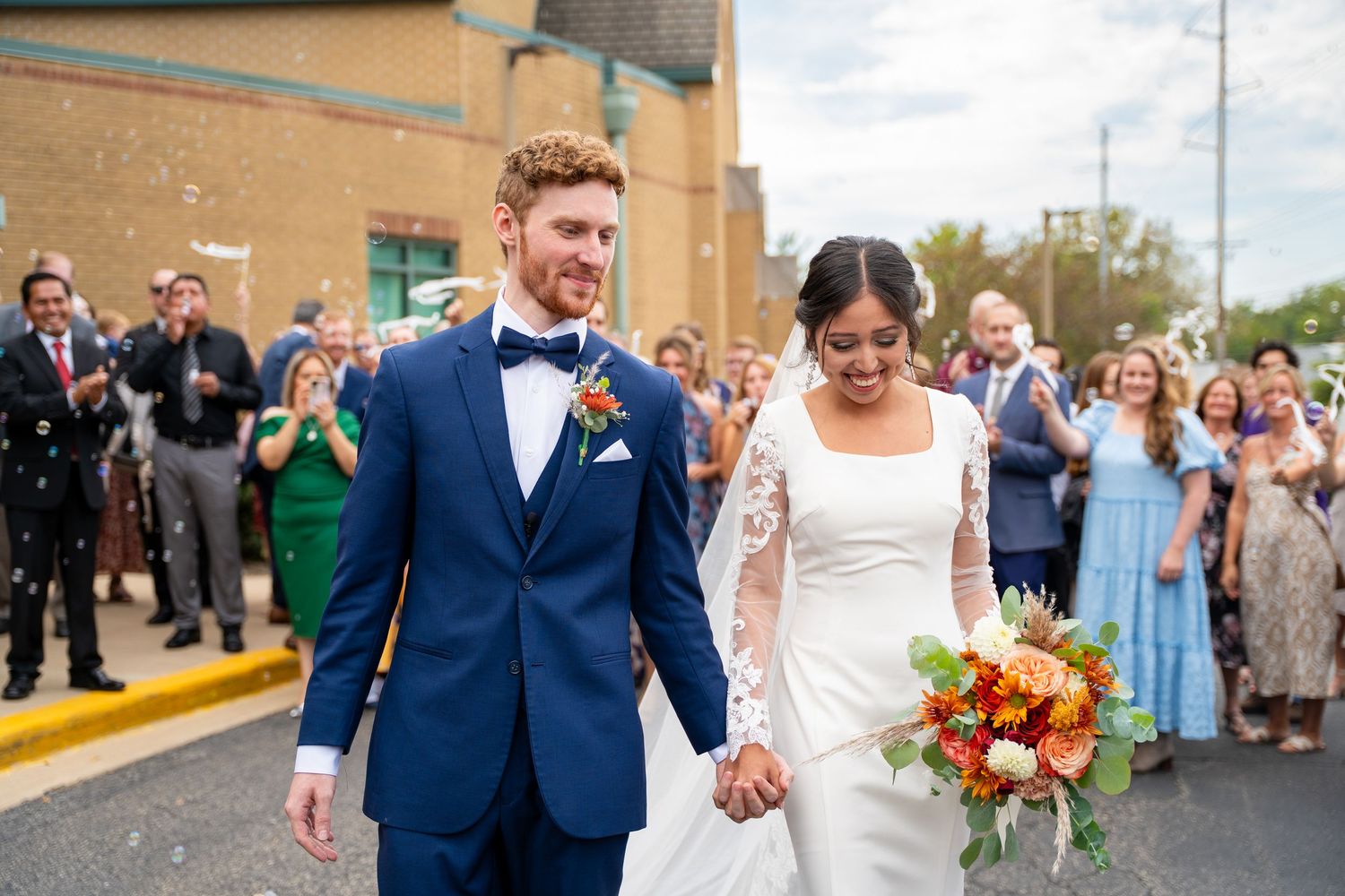 A newlywed couple walks together while wedding guests cheer in the background on a sunny day.