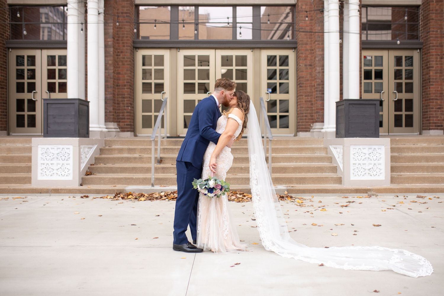 A couple shares an intimate moment on the steps of an elegant building with white columns and windows.