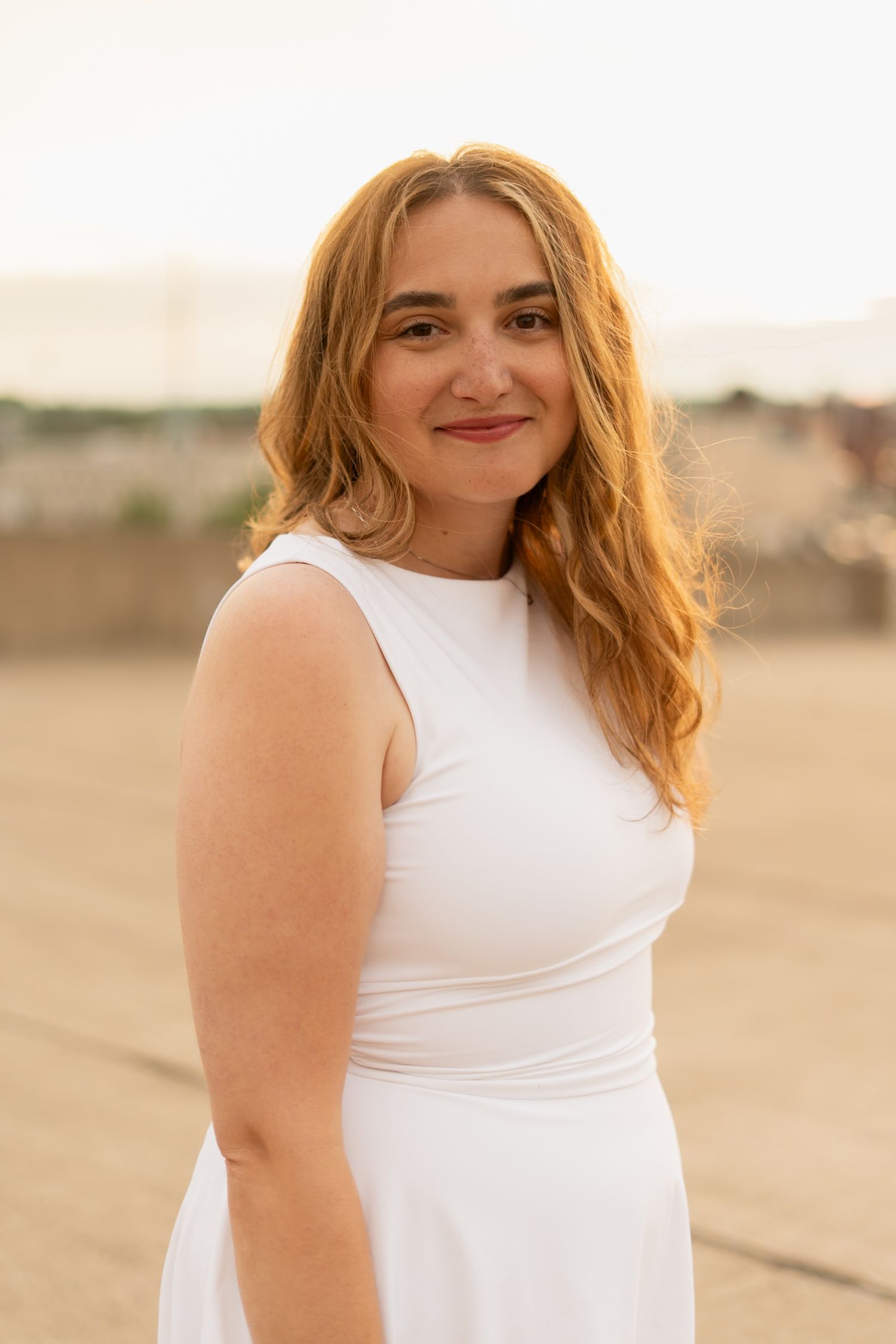 Woman in white sleeveless top poses against warm sunset backdrop.