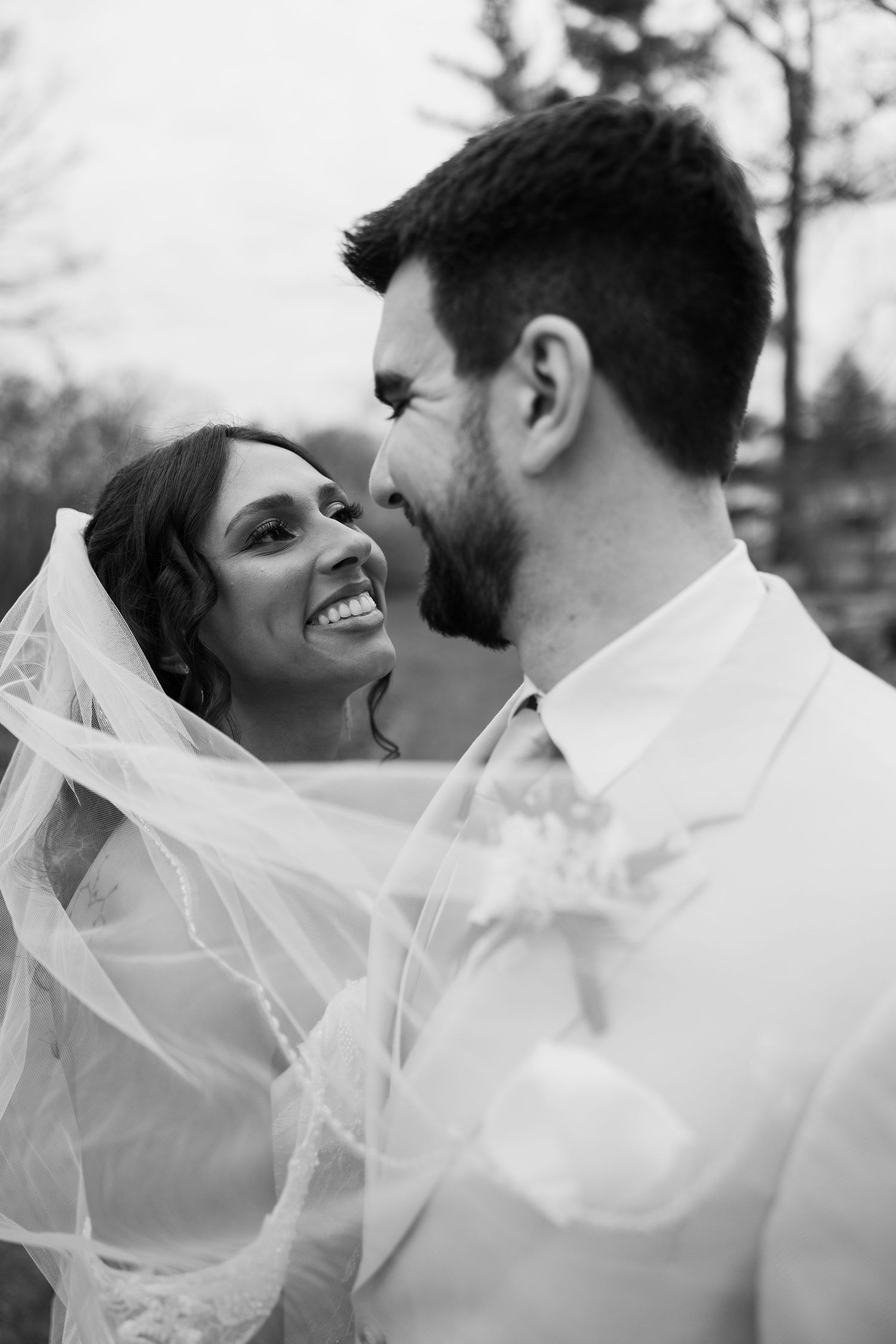 Black and white wedding photo of smiling bride in veil gazing at bearded groom outdoors.
