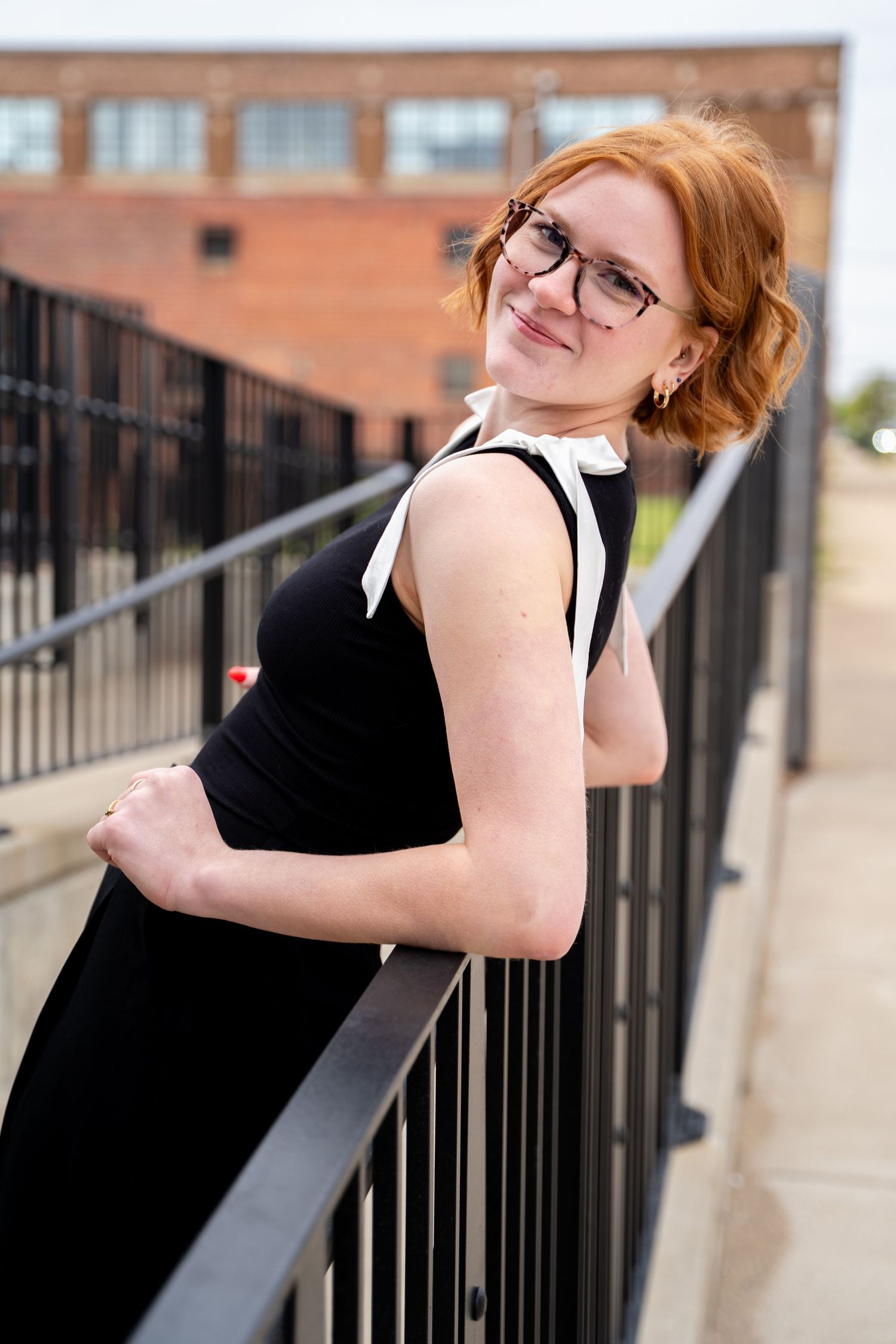Woman in black dress poses playfully against metal railing in urban setting.