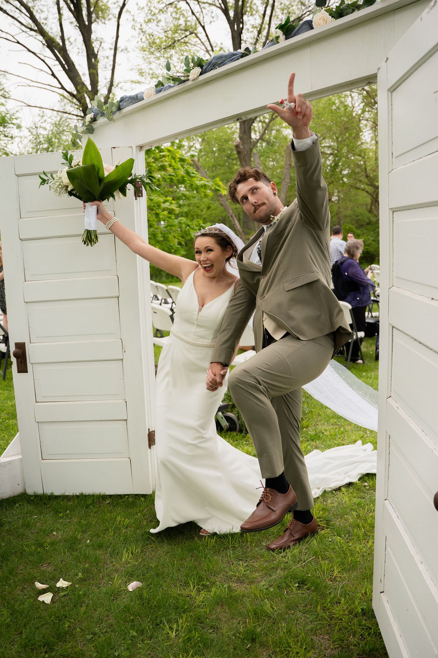 Joyful newlyweds celebrate exiting ceremony through white door arch, bride holding bouquet, groom giving peace sign.