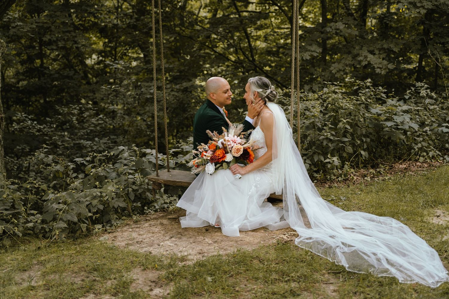 Bride and groom sitting on a wooden swing surrounded by lush greenery, bride holding a colorful bouquet.