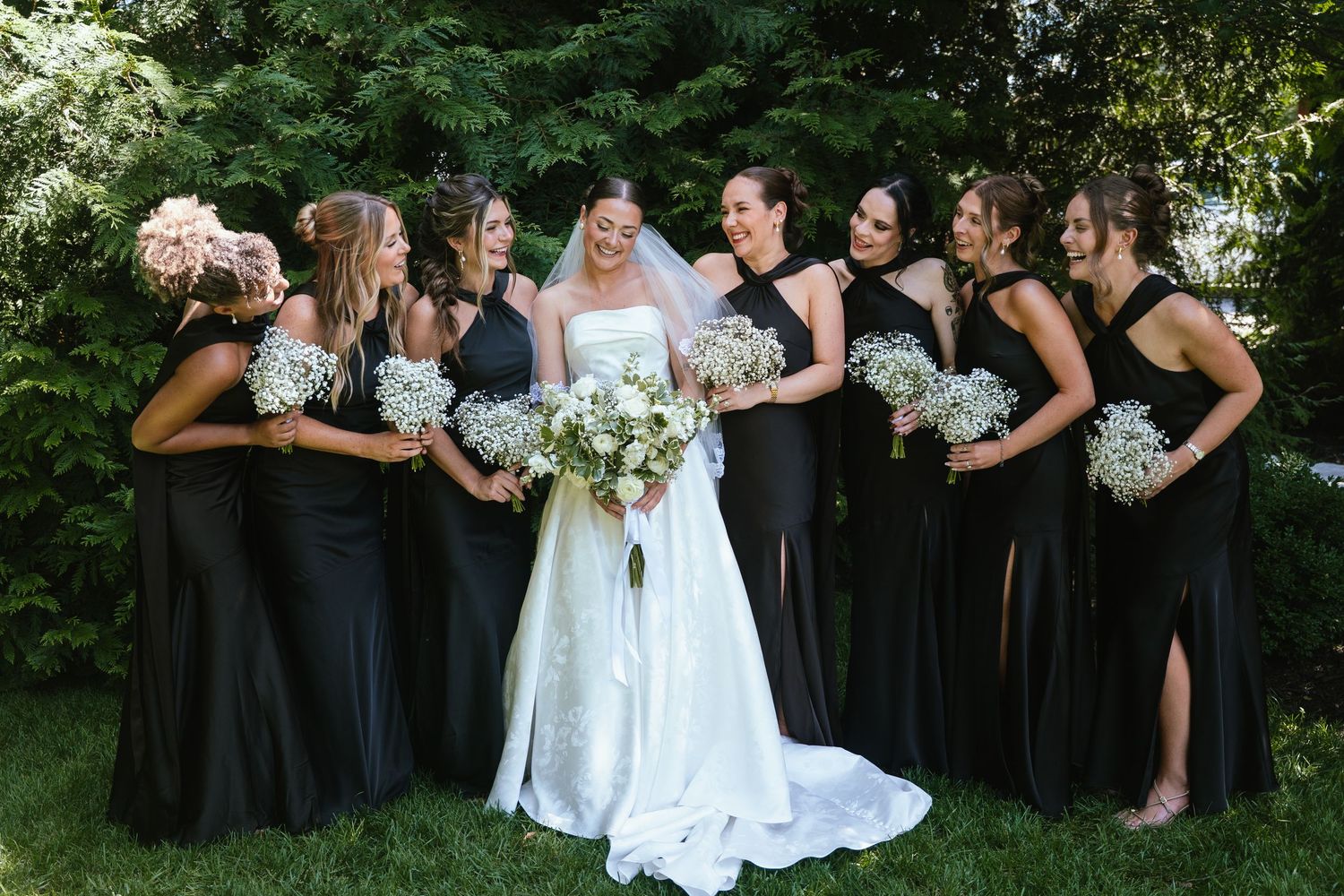 Bride in white gown with bridesmaids in black dresses holding white bouquets, smiling together outdoors.