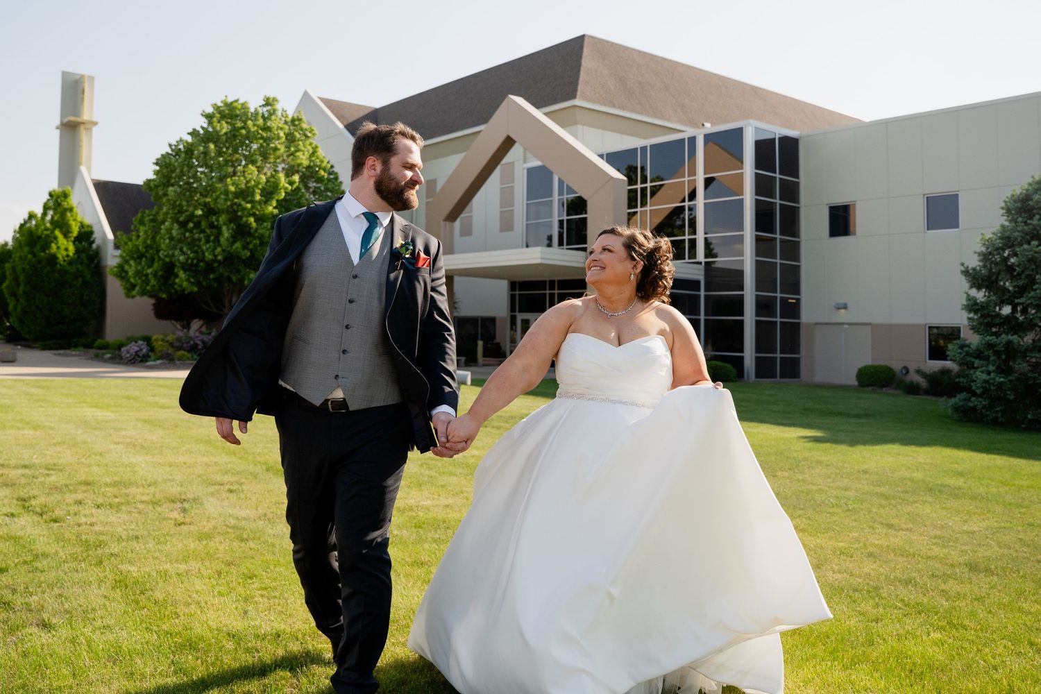 A couple in wedding attire spins and dances together on a sunny green lawn outside a white building.