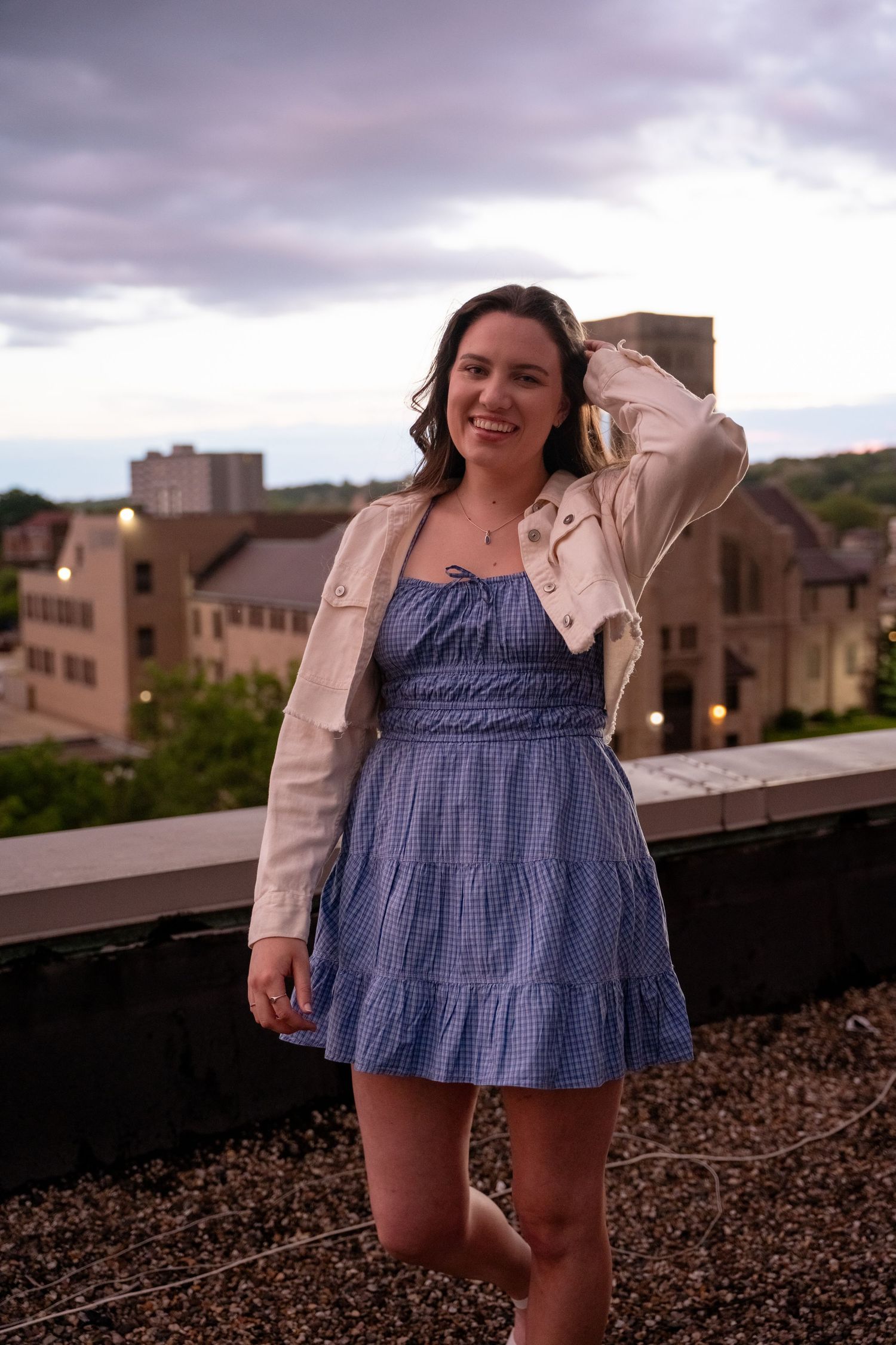 Woman in blue dress poses playfully on a rooftop with urban buildings in background.