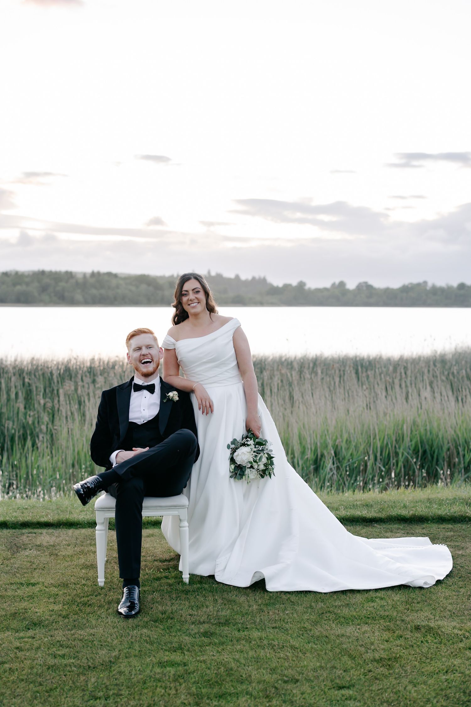 Bride in white gown holds bouquet beside groom in black tuxedo seated on chair, with lake and reeds backdrop.