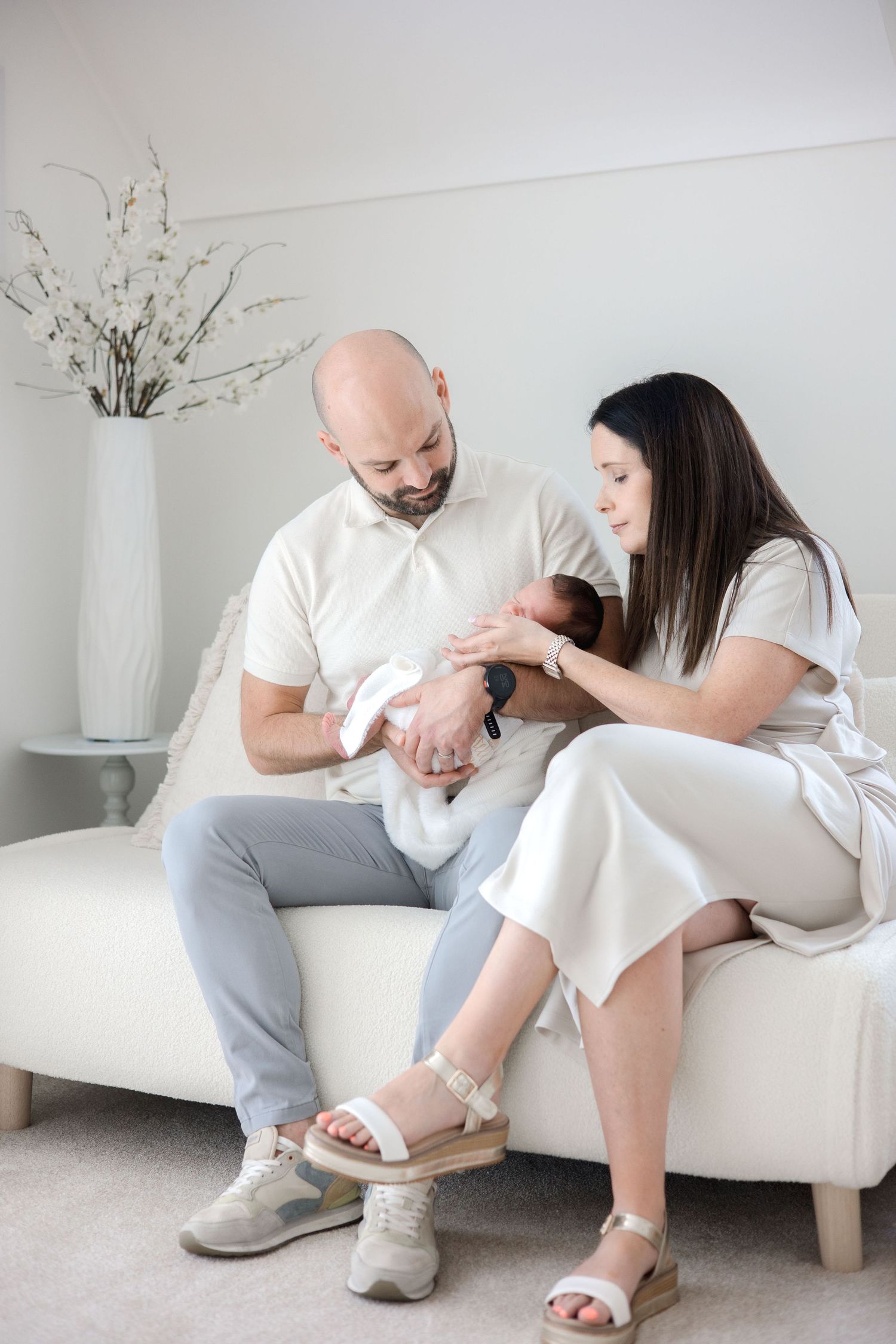 Parents sitting on a white couch lovingly gazing at their newborn baby in a bright, minimalist room.