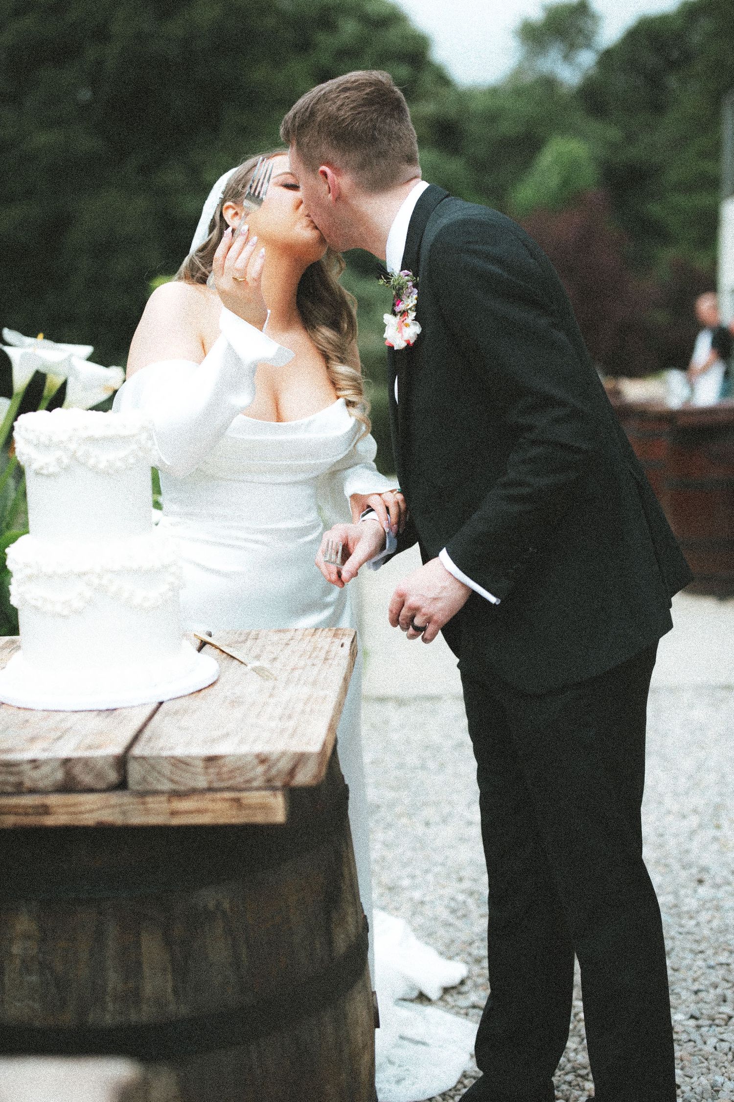 Bride and groom share a kiss while cutting their white wedding cake outdoors at a rustic wooden table.