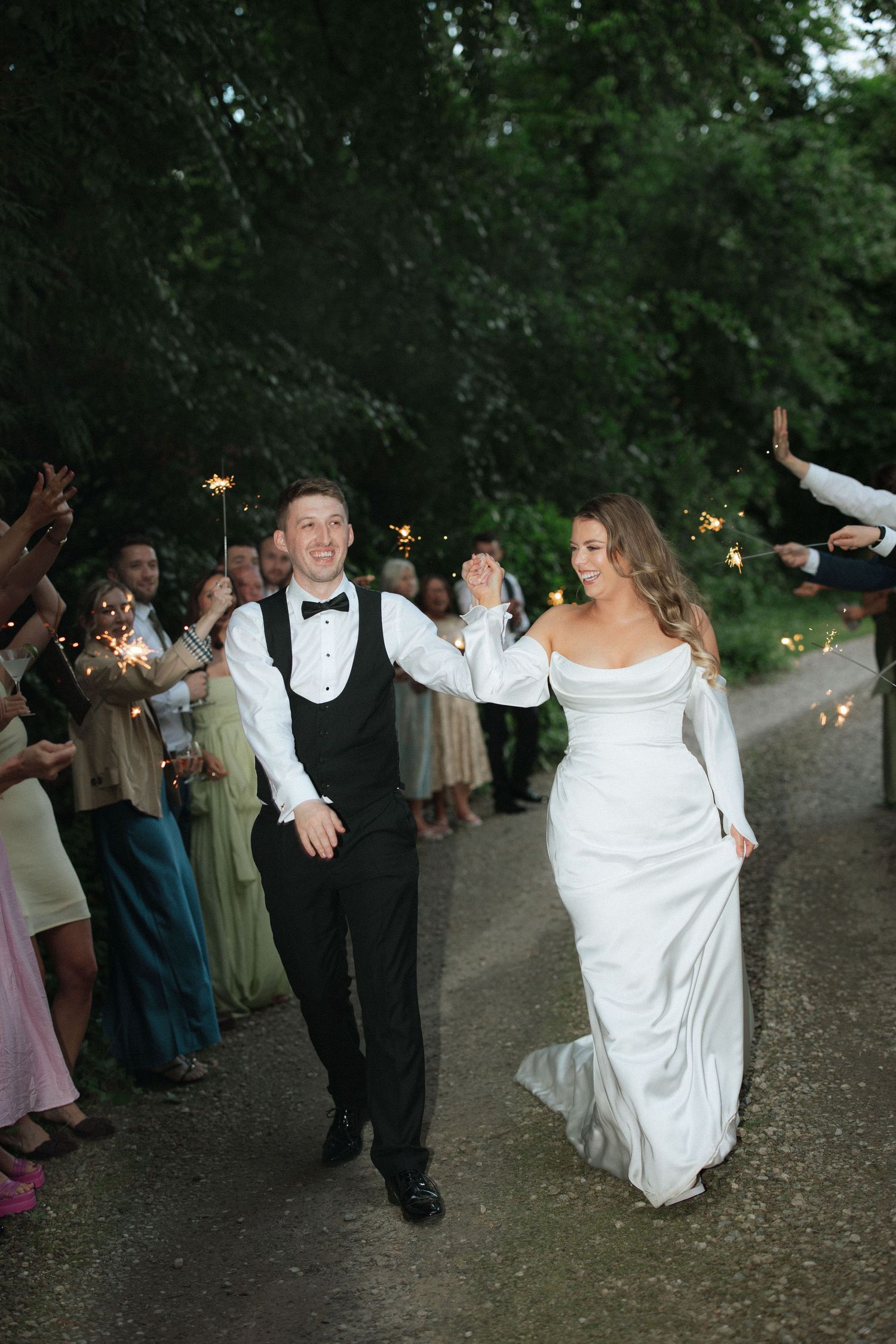 Bride and groom walk through sparkler exit lined with guests at outdoor evening wedding celebration.