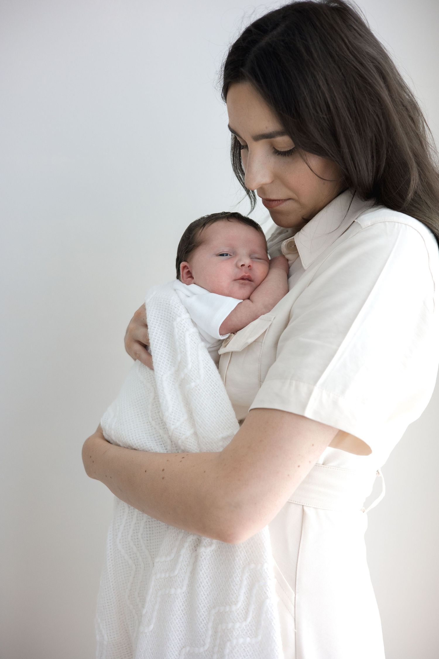 Mother in white dress gently holding swaddled newborn baby against a light background.
