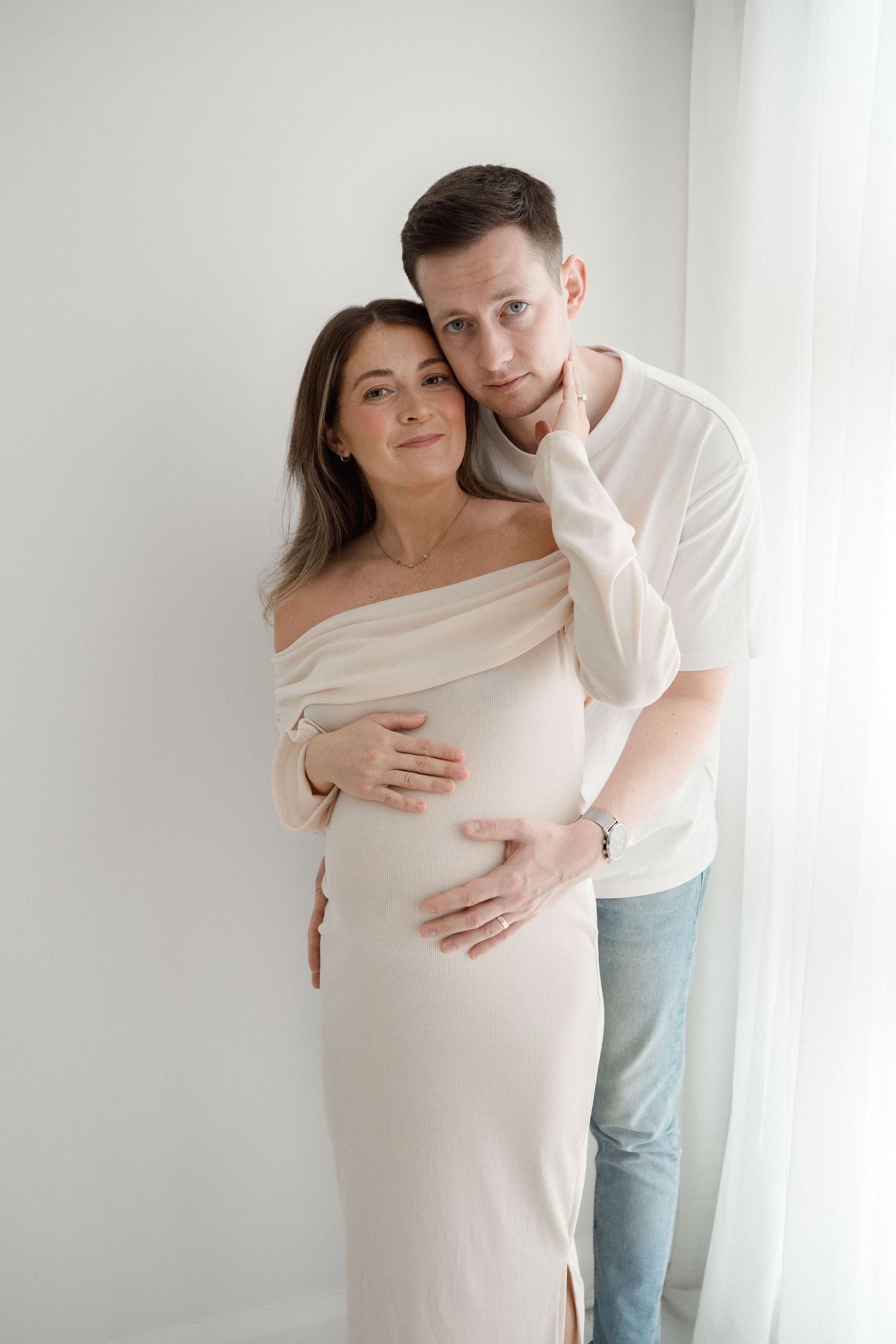Couple posing in a bright studio, man embracing pregnant woman in elegant cream ribbed maternity dress.