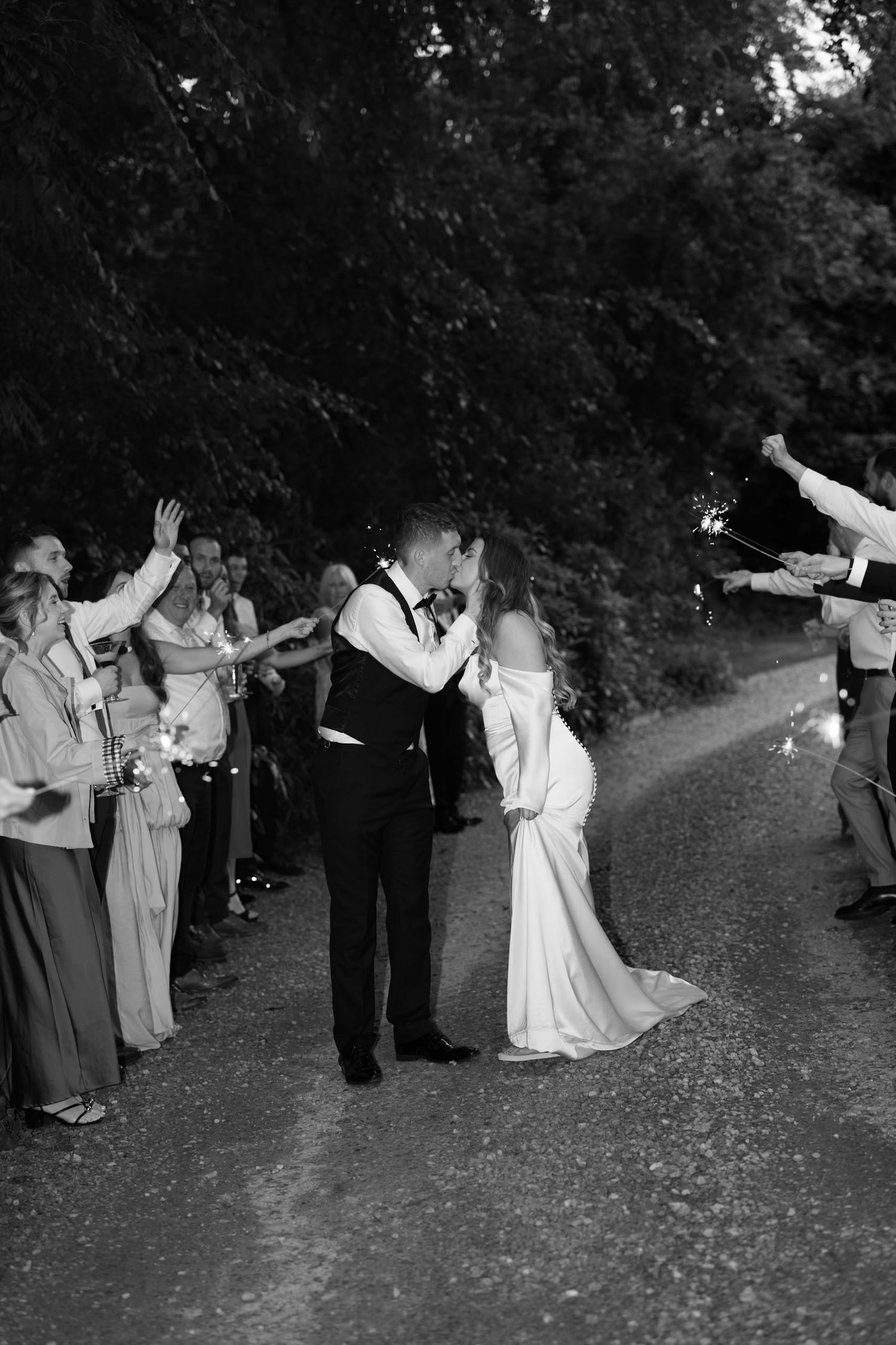 Black and white photo of newlyweds kissing during sparkler send-off lined with cheering wedding guests on a tree-lined path.
