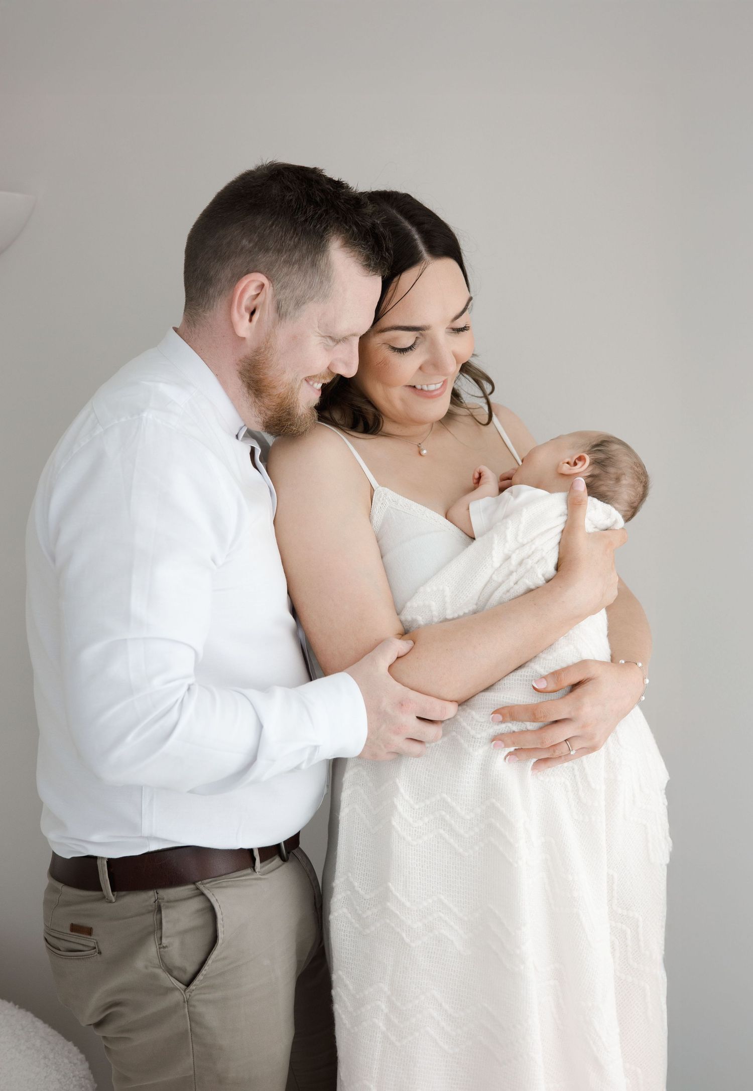 Smiling couple in white outfits standing close together holding their newborn baby.