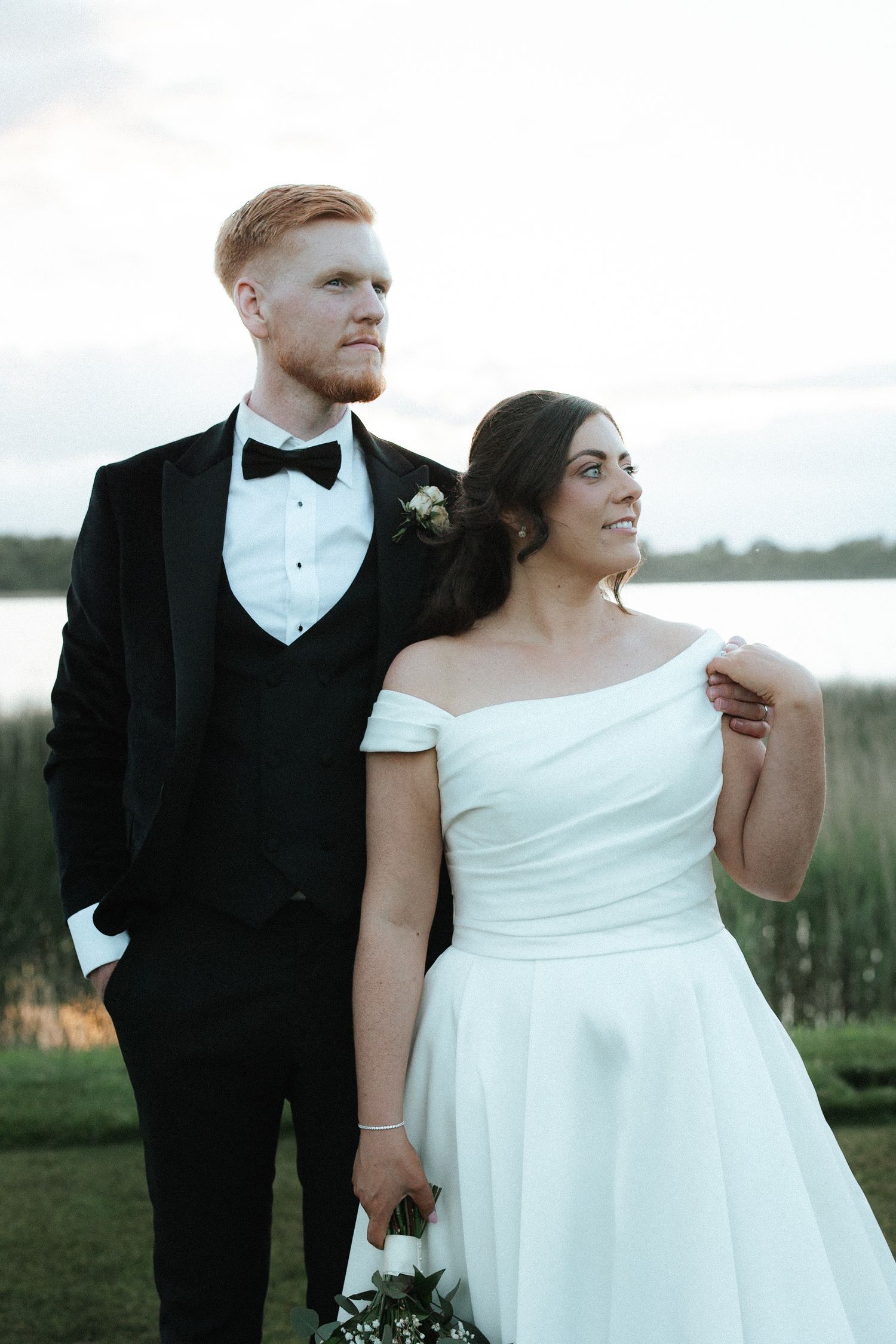 Bride and groom posing elegantly outdoors by a lake in wedding attire at dusk.