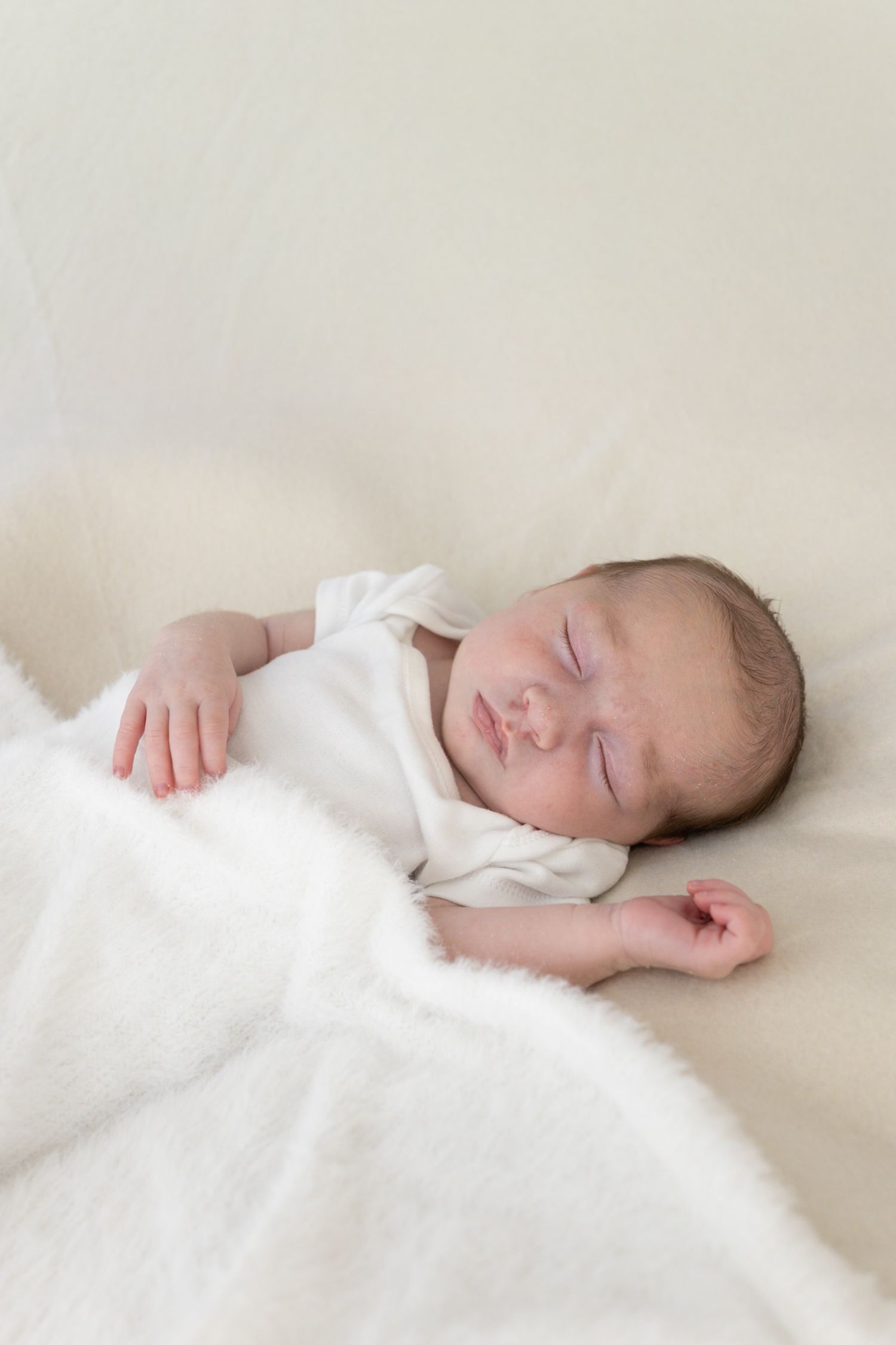 Sleeping newborn baby dressed in white onesie resting peacefully on a soft cream fluffy blanket.