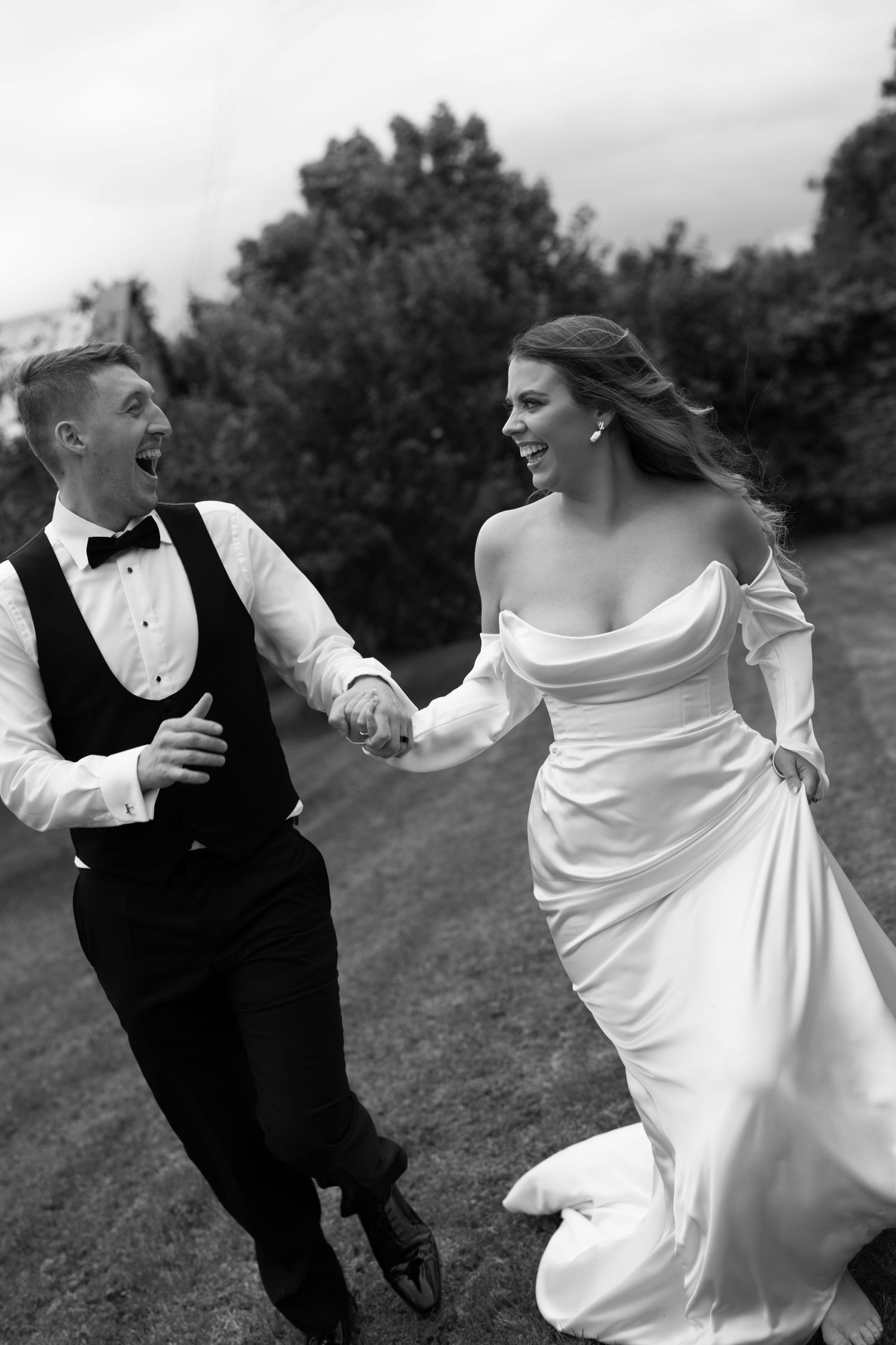 Black and white photo of a joyful bride and groom holding hands and running outdoors on their wedding day.