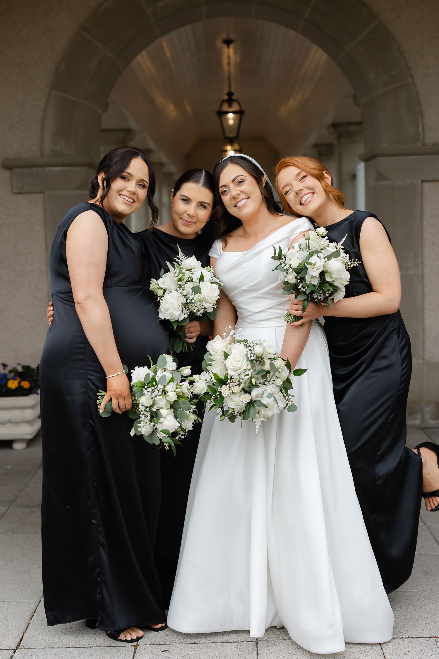 Bride in white gown poses with three bridesmaids in black dresses holding white floral bouquets outside elegant venue.