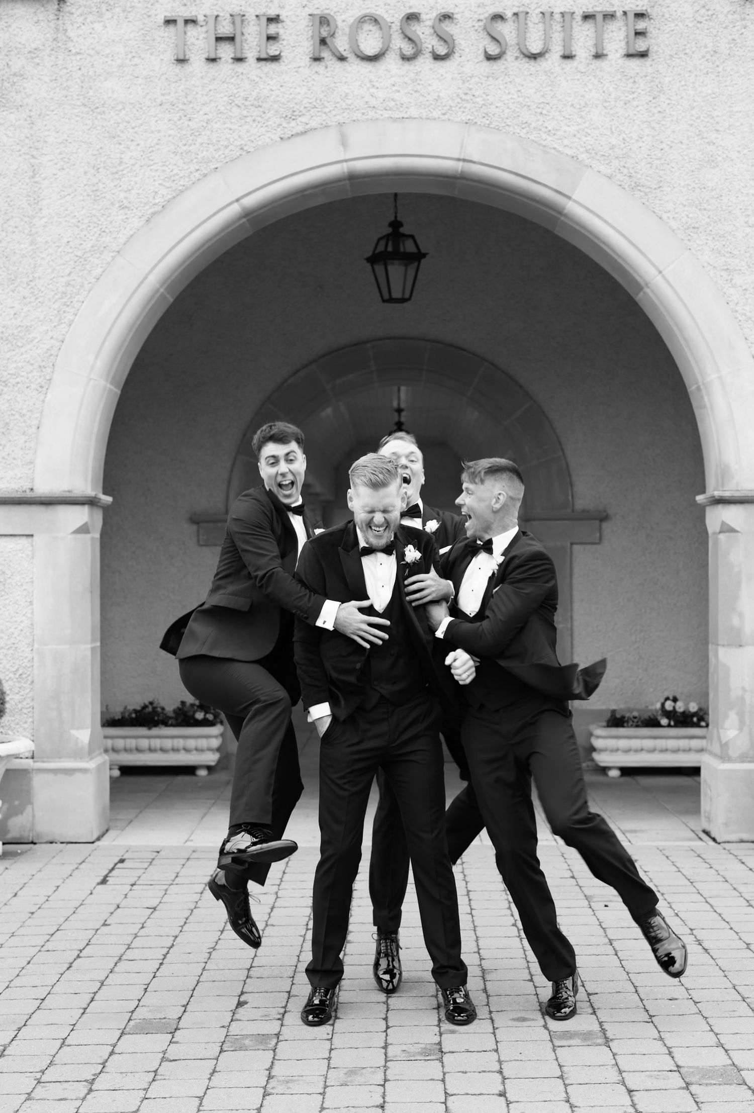 Groomsmen in tuxedos celebrate joyfully outside The Ross Suite in a black and white wedding photo.