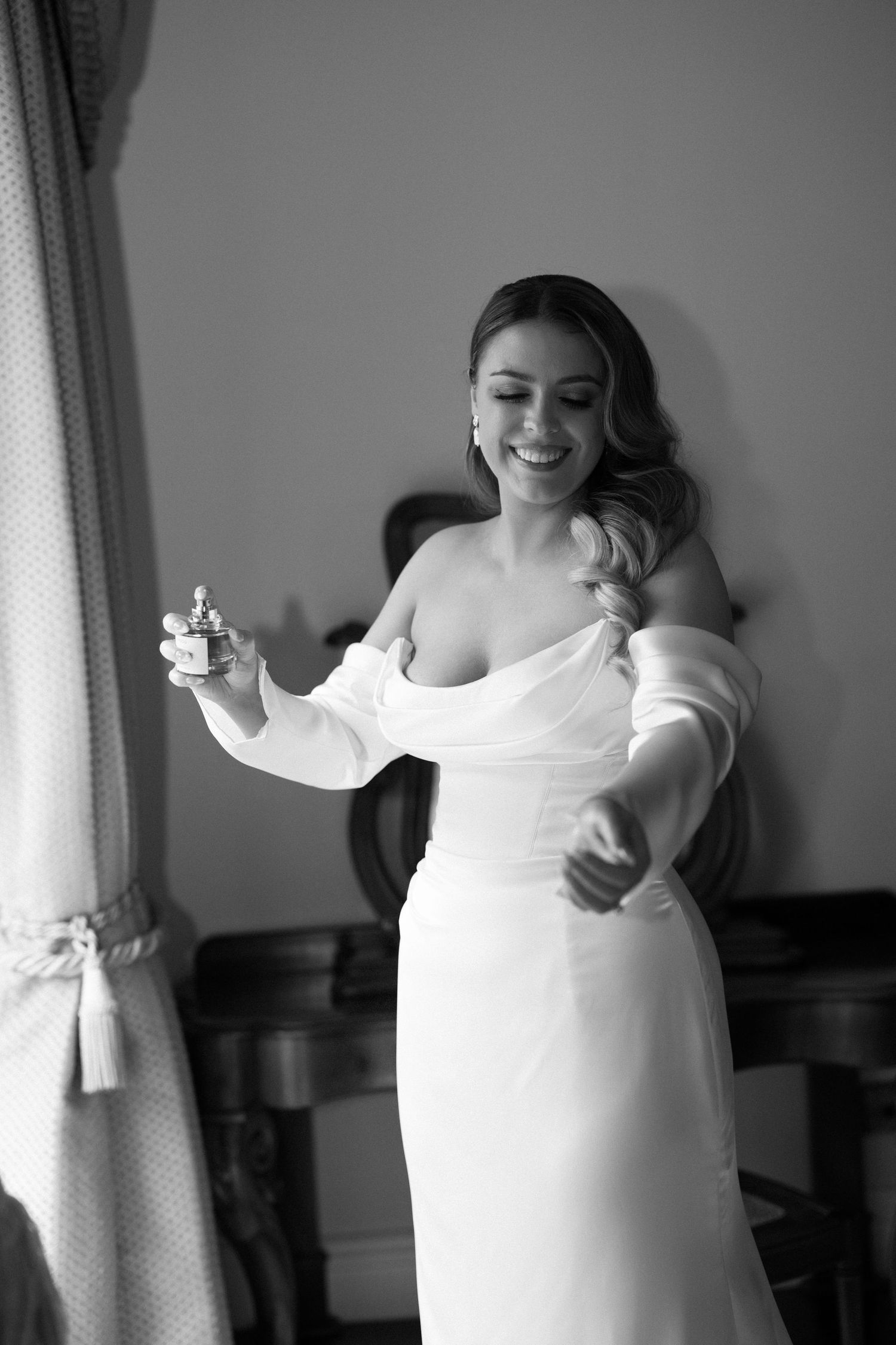 Smiling bride in elegant white off-shoulder gown applying perfume while getting ready on her wedding day.