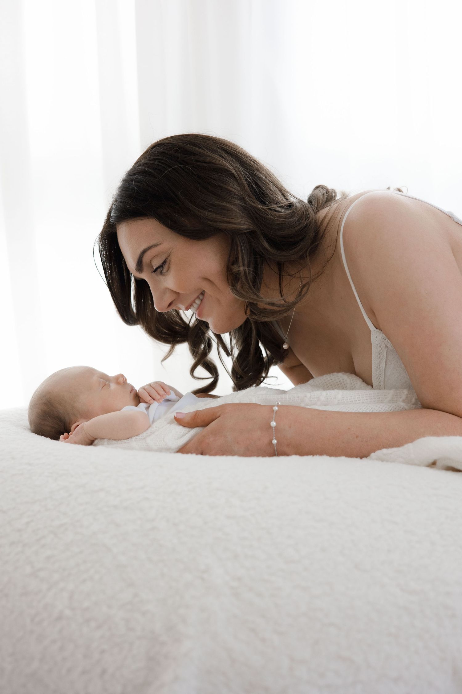 A smiling mother leans over her newborn baby lying on a fluffy white surface in bright light.