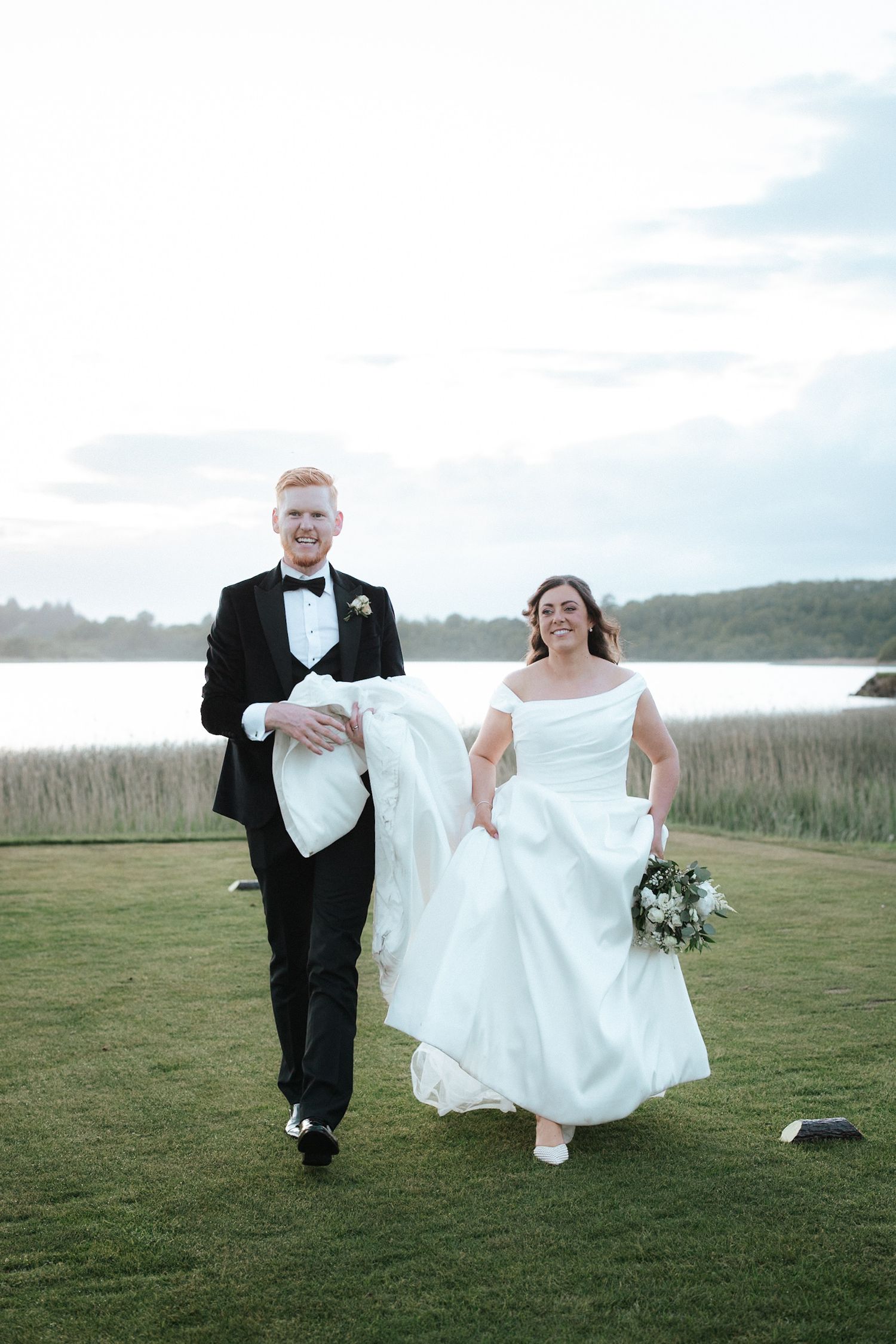 Smiling bride and groom walking hand in hand on green lawn near waterfront, bride holding bouquet in white off-shoulder gown.