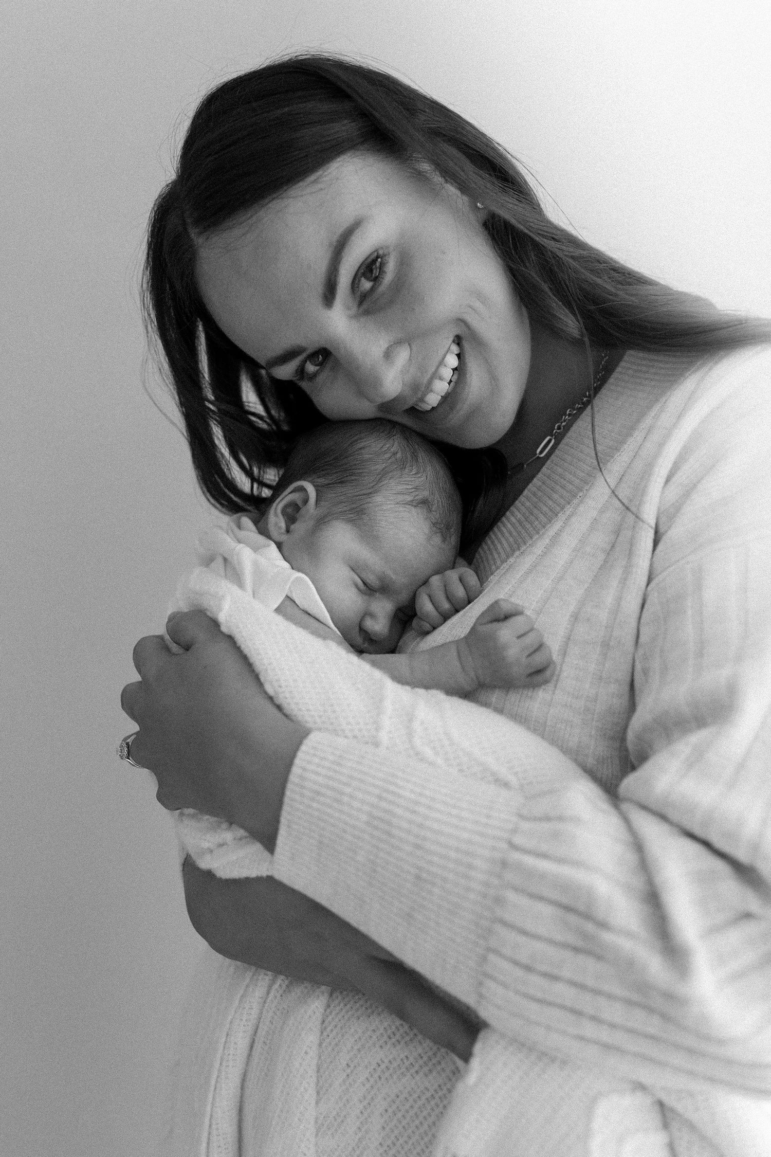 Smiling mother in white robe holding sleeping newborn baby in black and white portrait.