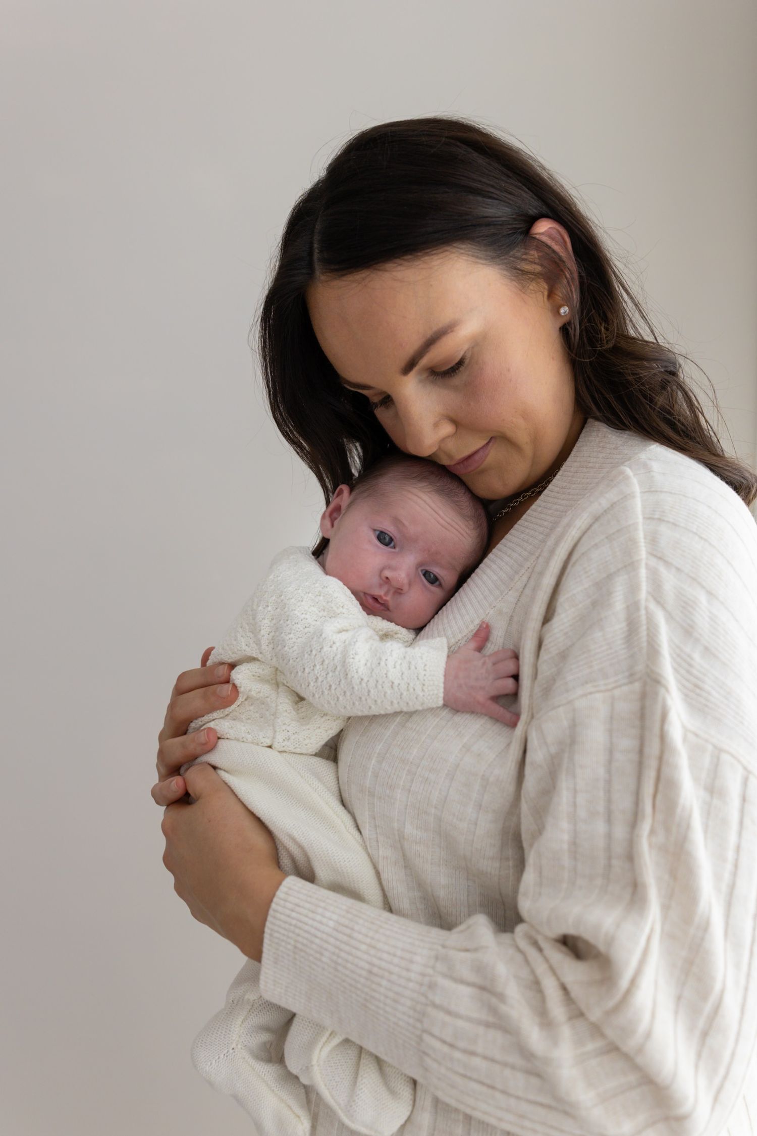 Mother in cream sweater gently cradling her wide-eyed newborn baby.